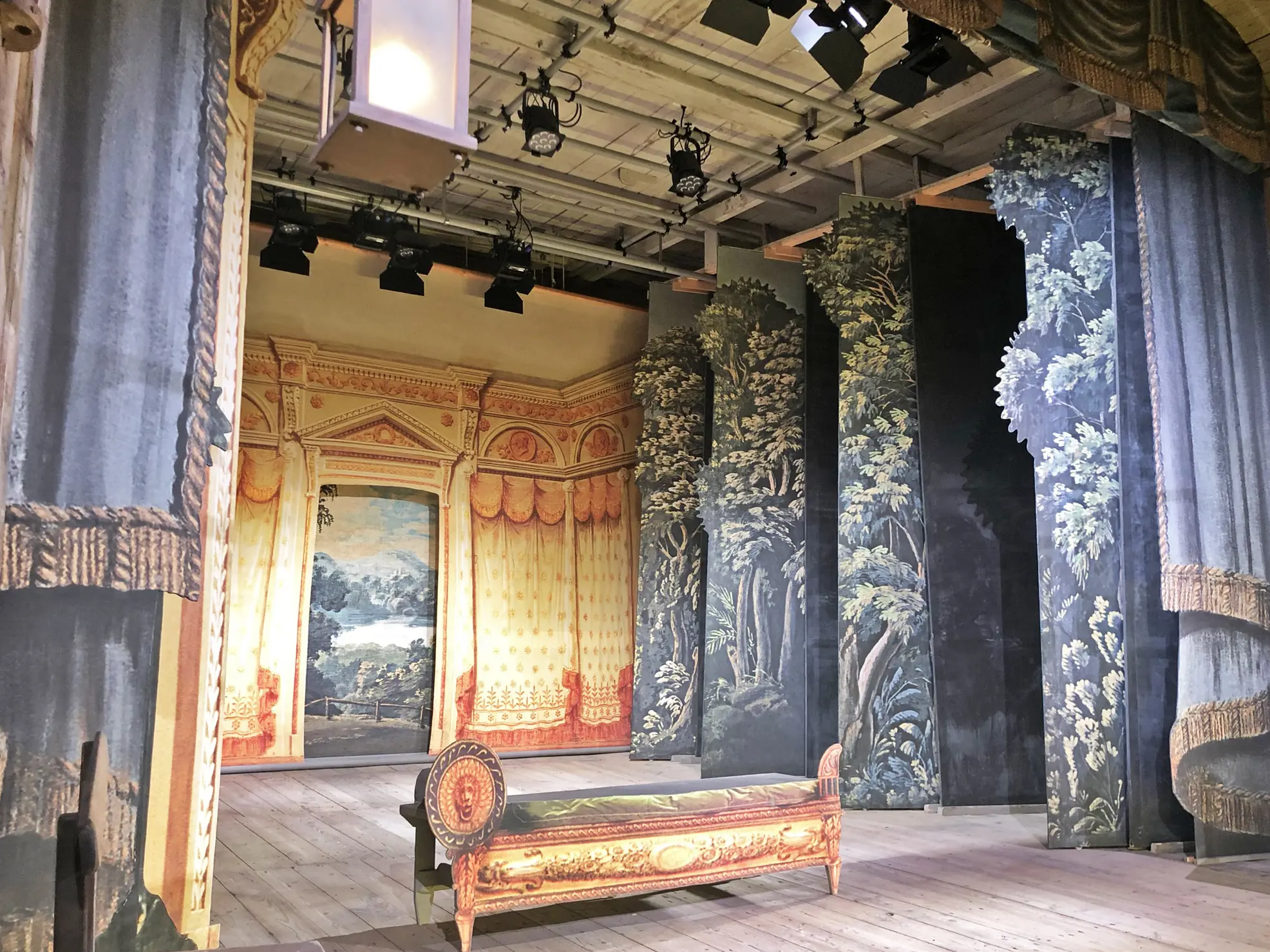 Vadstena gamla teater. Scenen har plankgolv och kulisser som föreställer draperier och växtlighet. I bjälktaket hänger strålkastare.