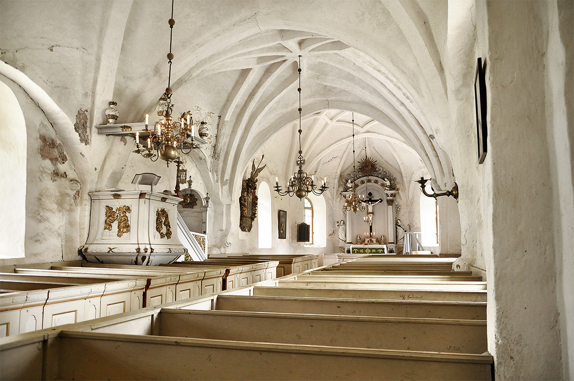 Ett kyrkorum med ljust putsad fasad och välvda tak med träbänkar som utgör altargången.