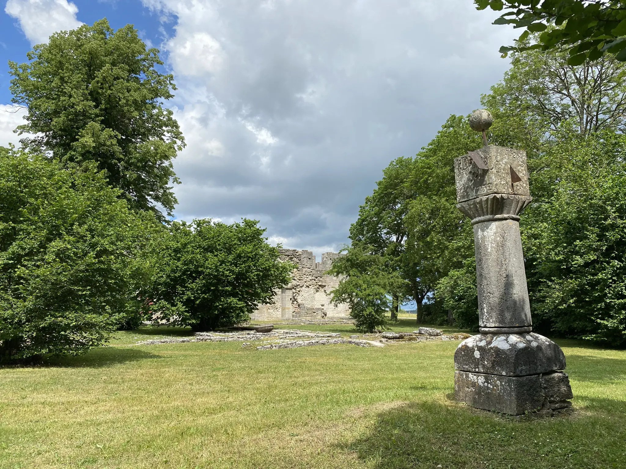 Parken vid Roma kloster. Kyrkoruinen och de gamla stengrunderna omges av lummiga träd. I förgrunden en stenskulptur som liknar en medeltida pelare.