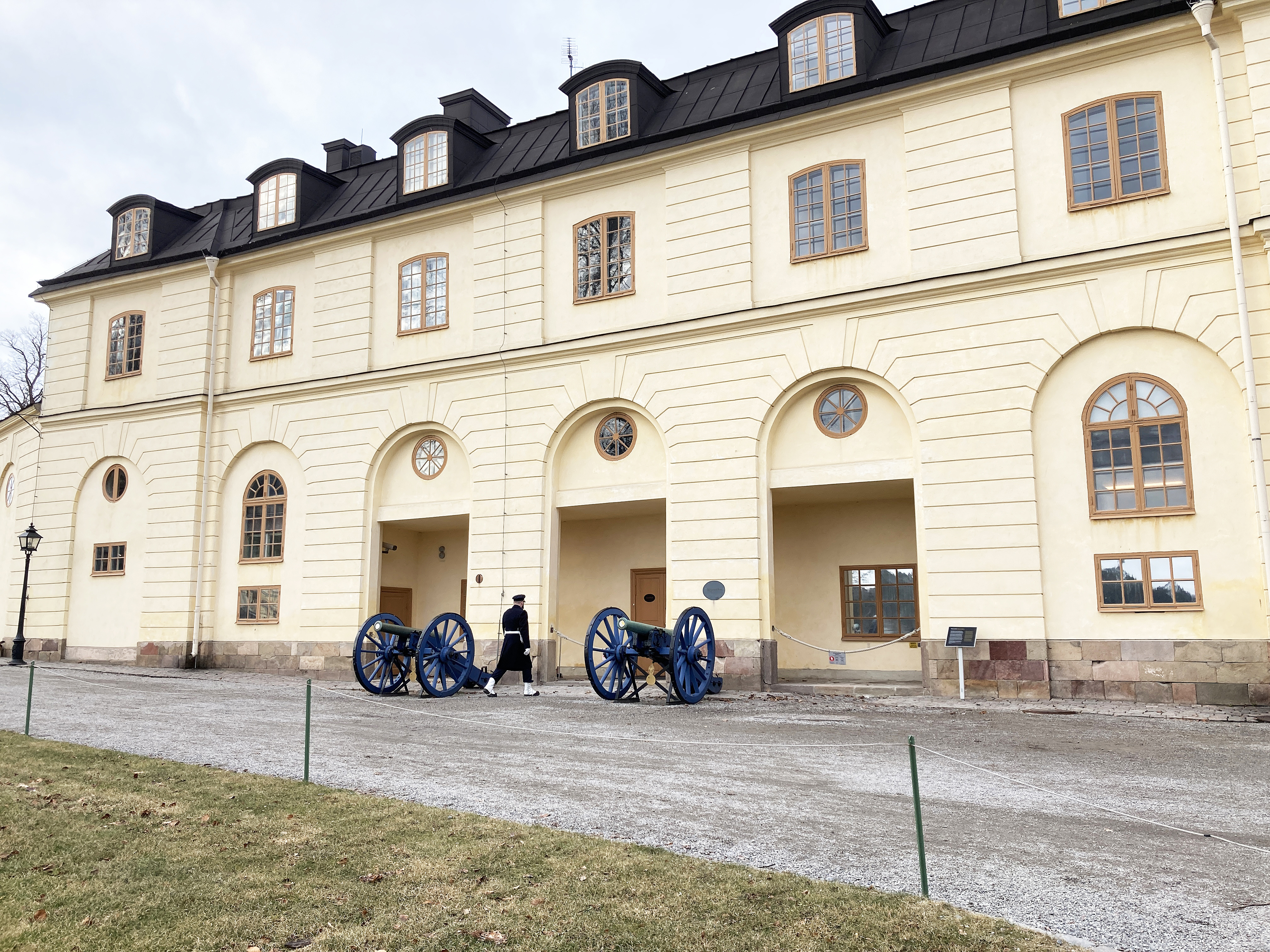 Drottningholms slotts-stall. Framför den gula byggnaden står två blå, plomberade kanoner. En vakt i vita damasker är på väg in i huset.