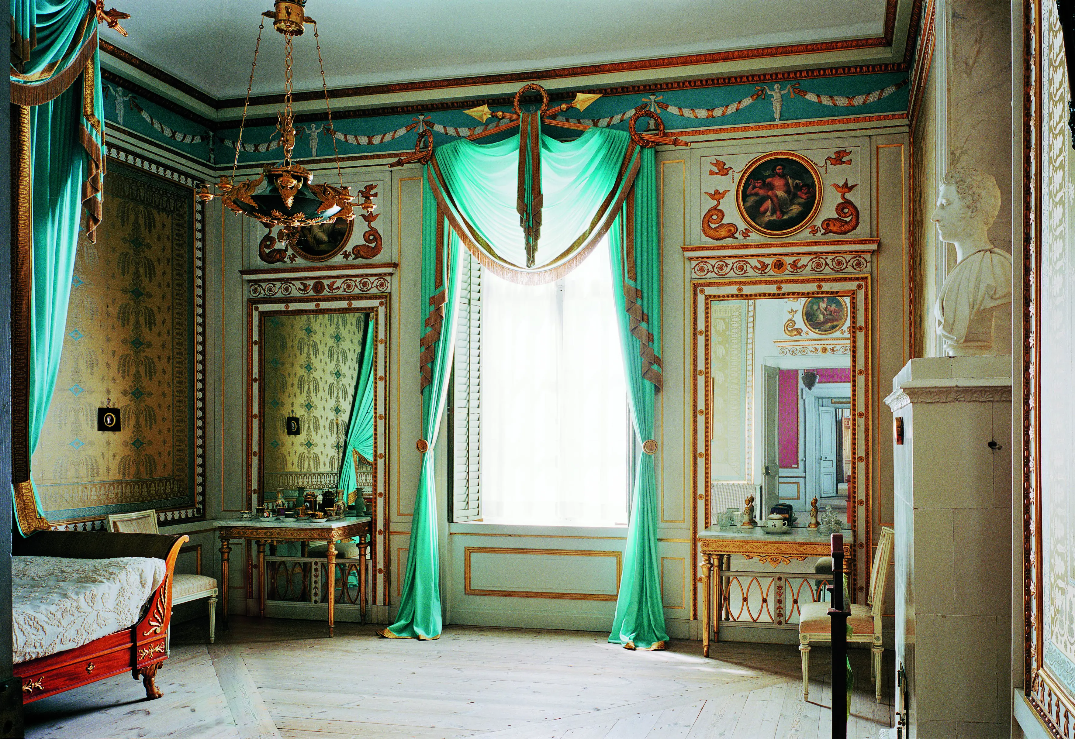 Ett sovrum i Rosersbergs slott med ett stort fönster mitt i rummet där solen skiner in. Fönstret har ljusgröna gardiner och inredningen är äldre med dekorerade tapeter och väggar.