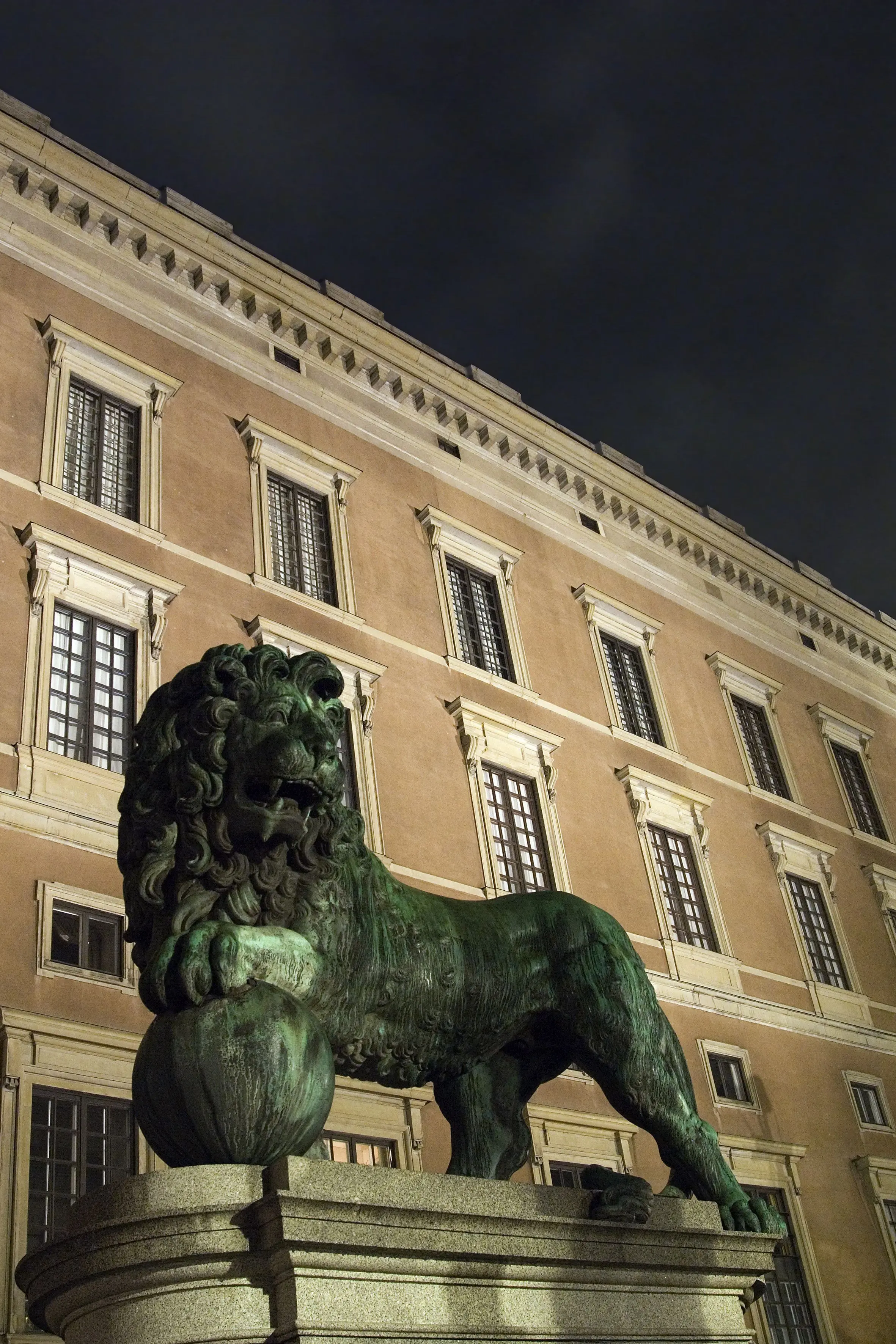 Stockholms slott. Del av fasaden i kvällsbelysning. I förgrunden en brons-staty av ett lejon med tassen på ett klot .