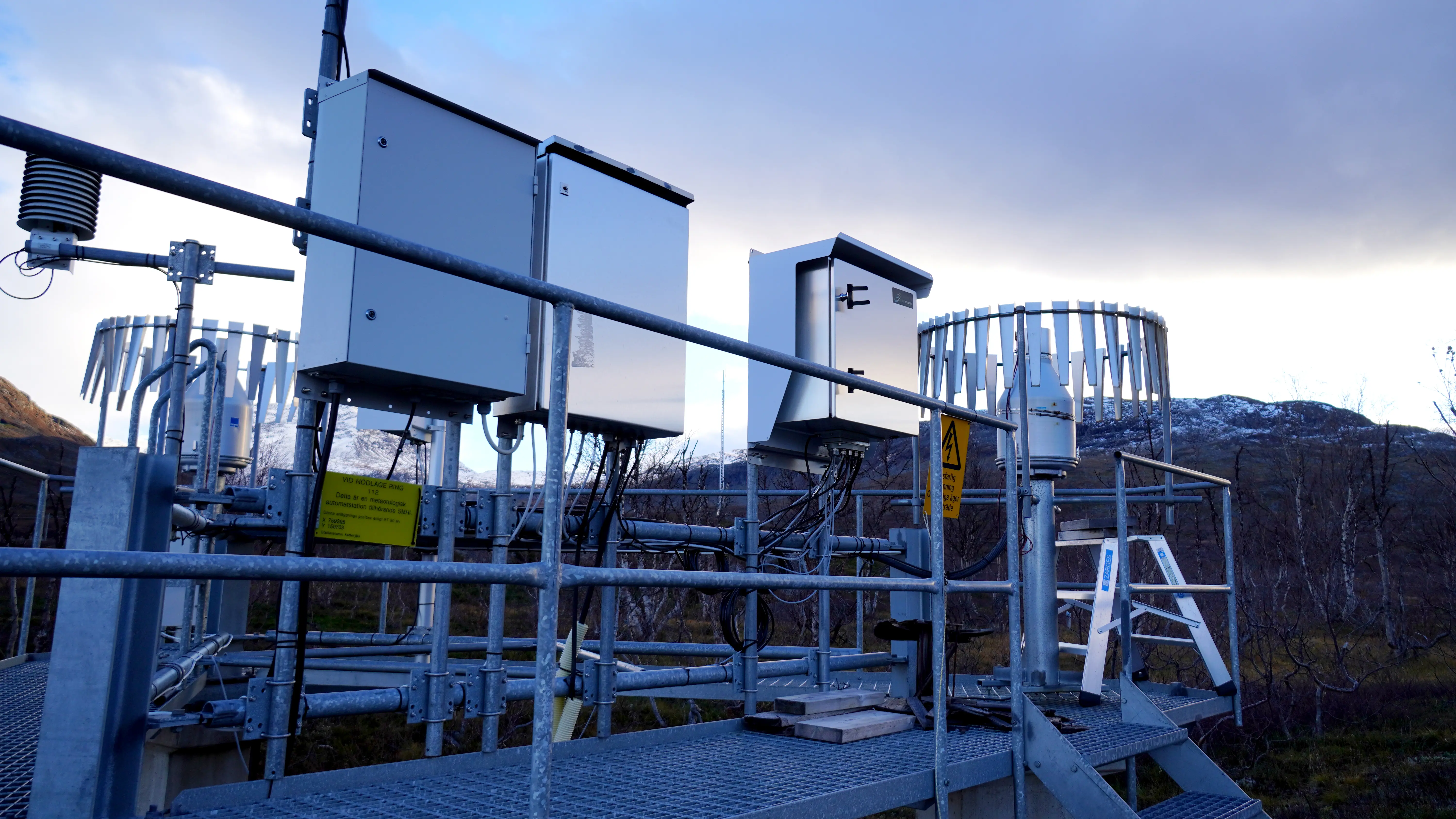 En väderstation som består av ett antal mätinstrument i metall, mitt i ett fjällandskap i kvällsljus.