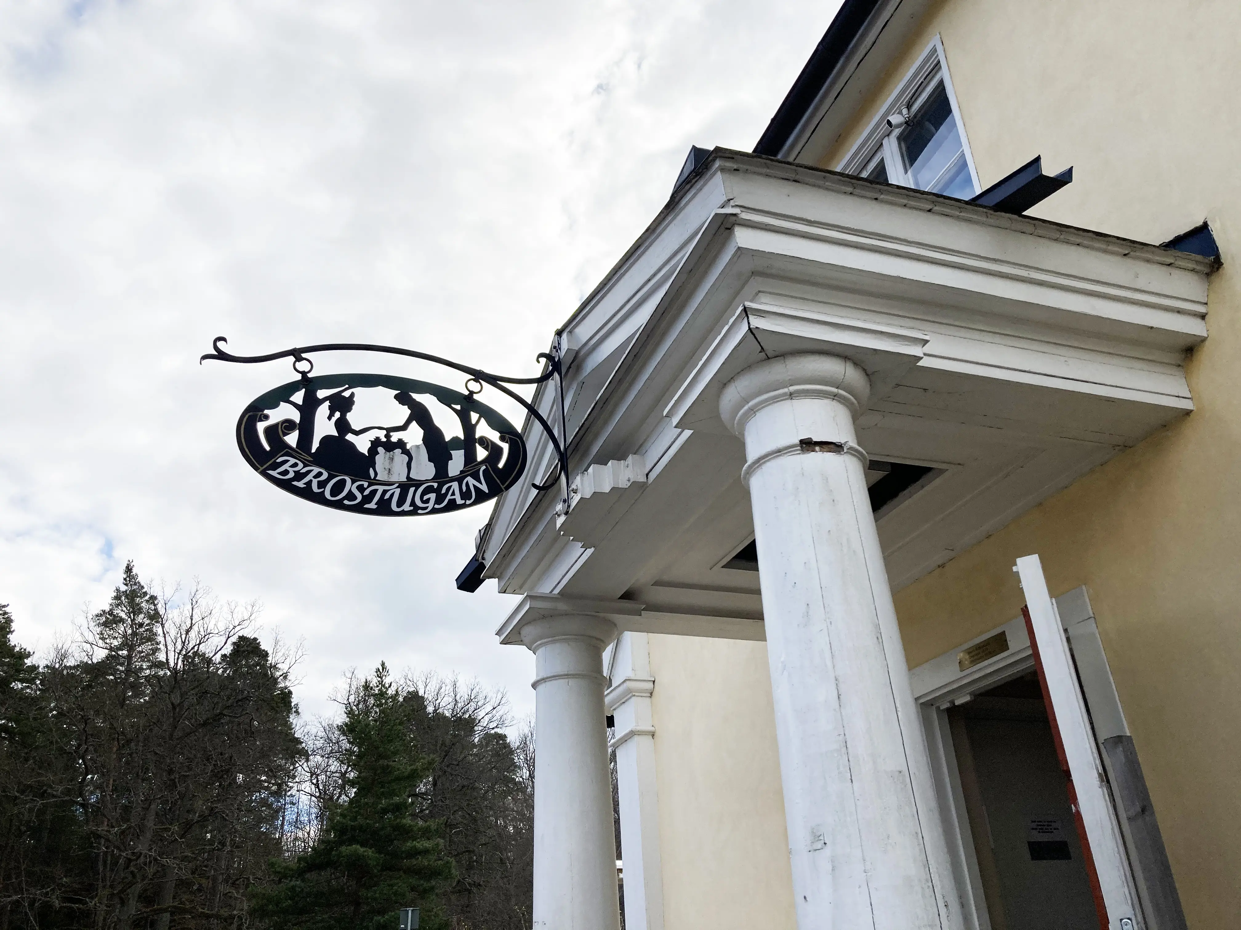 Brostugan på Kärsön. Entrén med klassicistisk portik i trä och en oval smides-skylt med "Brostugan" och siluetterna av två personer.