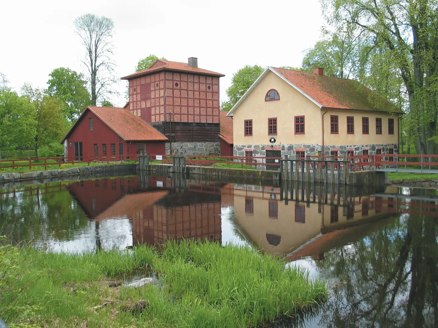 Tre hus omgivna av gröna träd som speglas på en vattenyta.