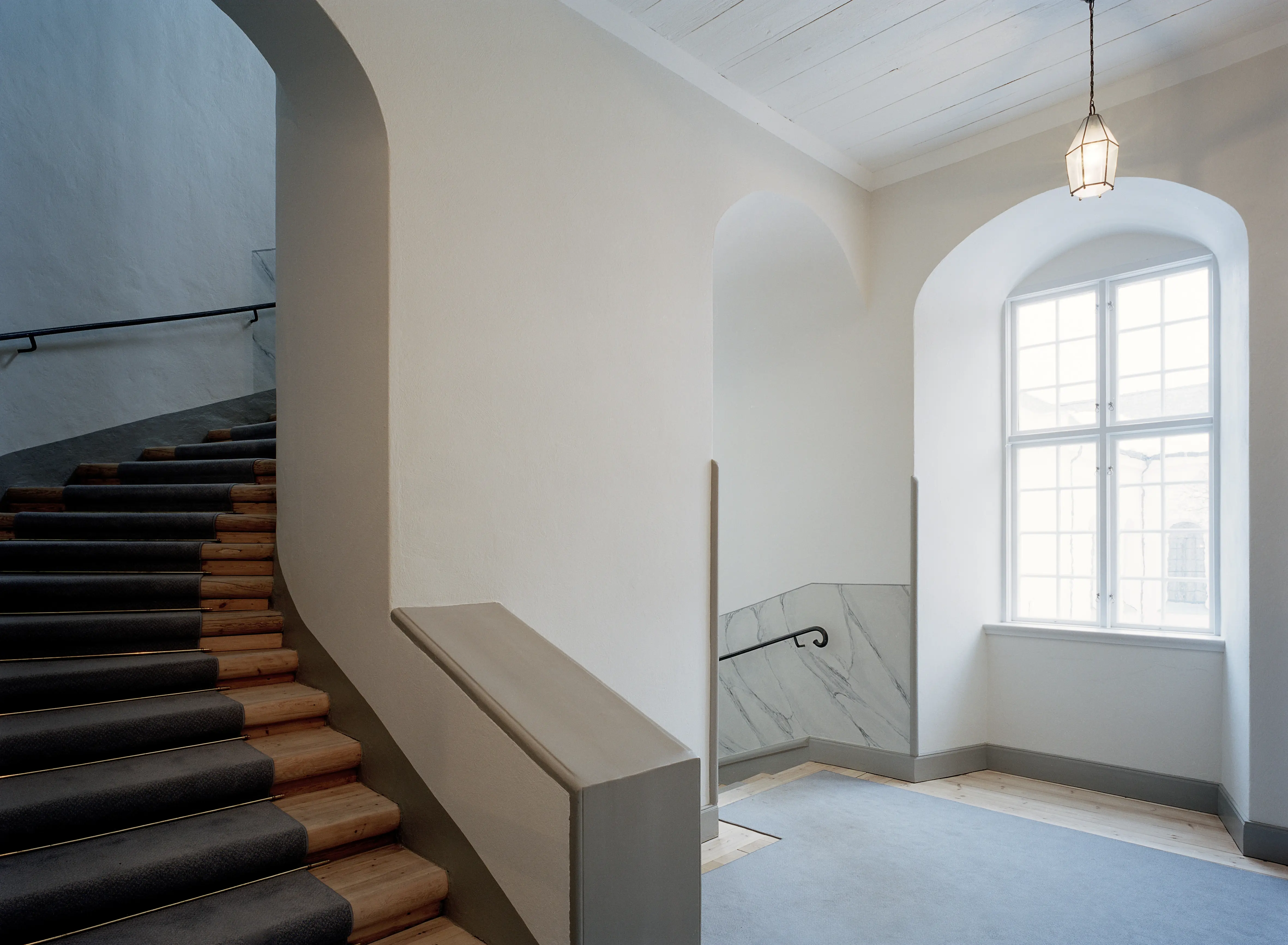 Trapphus i gamla gymnasiebyggnaden i Karlstad. Väggar och tak är vita. Ett stort, spröjsat fönster släpper in ljus.