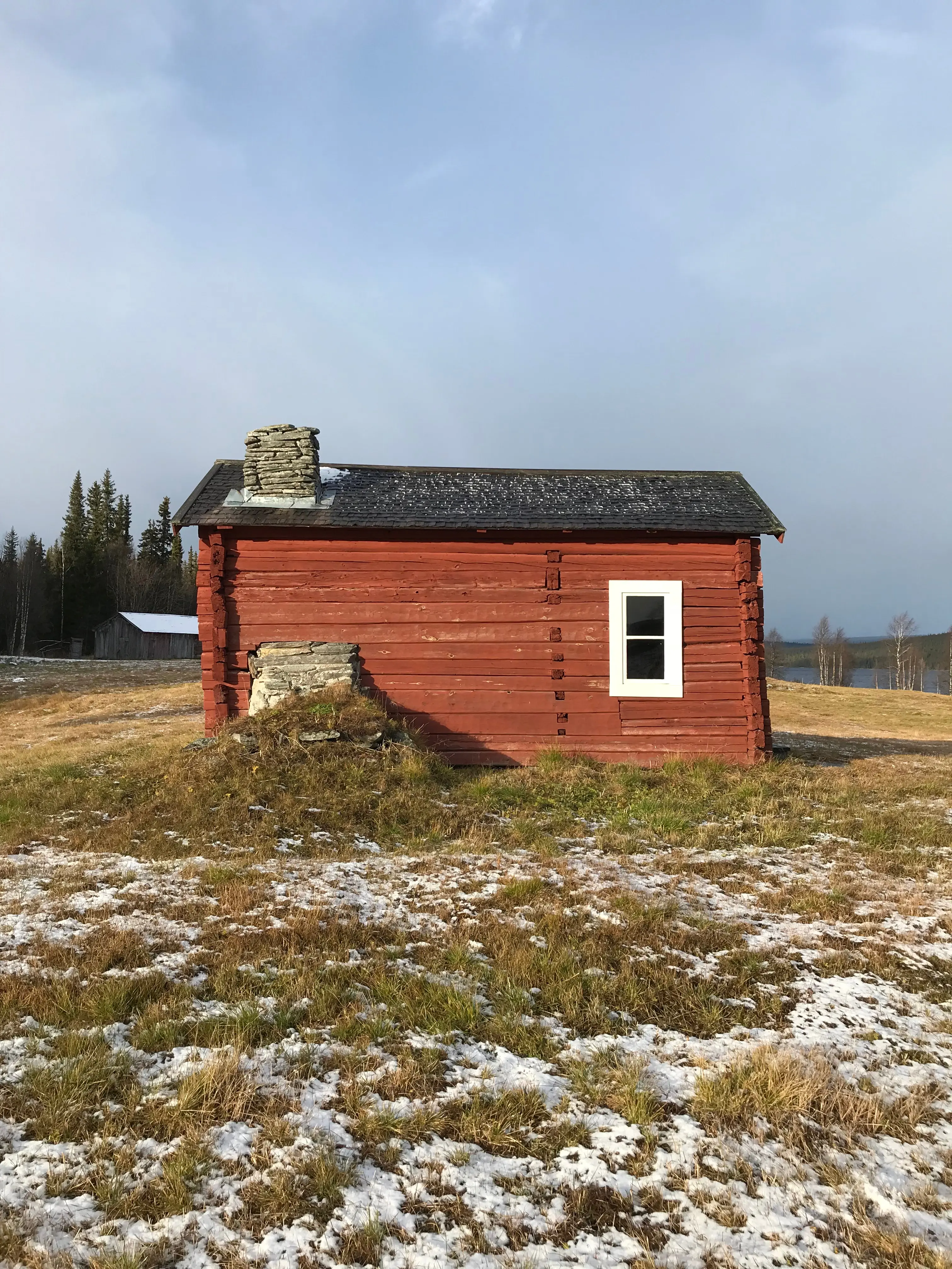 Åkernäs fjäll-lägenhet. En röd liten timmerstuga, med murstock och skorsten av sten. I bakgrunden skymtar sjön Överst-Juktan.