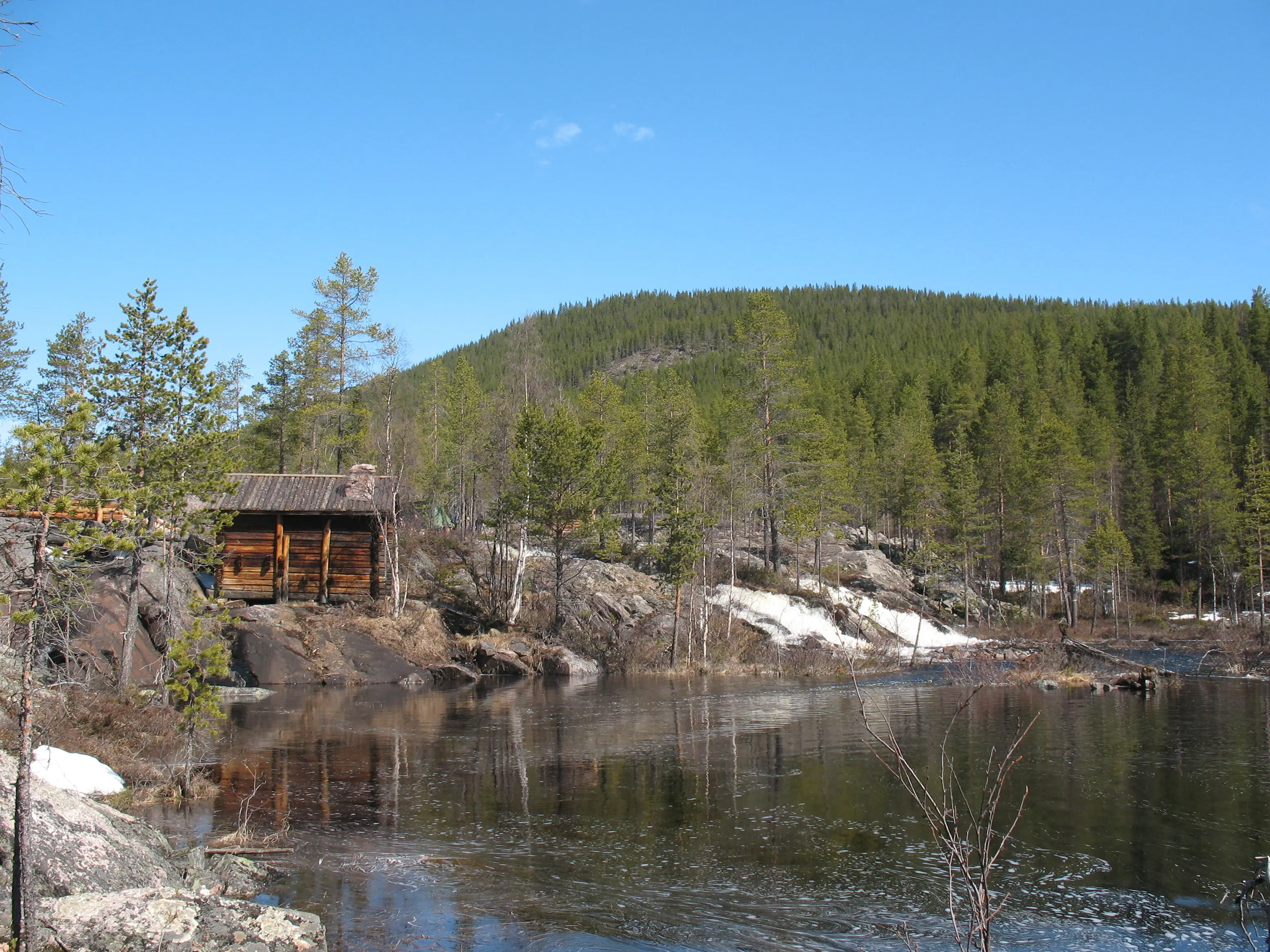 Granuddens skvaltkvarn på klipporna vid Kvarntjärn. I bakgrunden syns en bergsknalle med granskog. Vår och islossning.