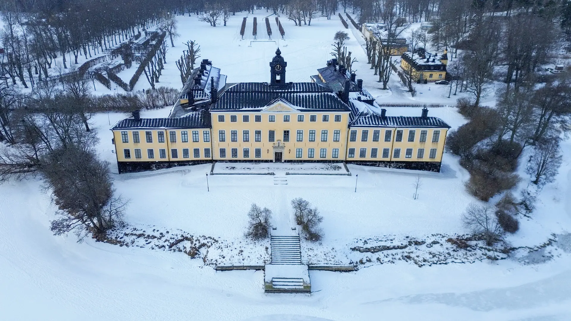 Drönarbild av Ulriksdals slott från vattnet. Framför slottet går en trappa ner till en brygga. Vinter och snö.