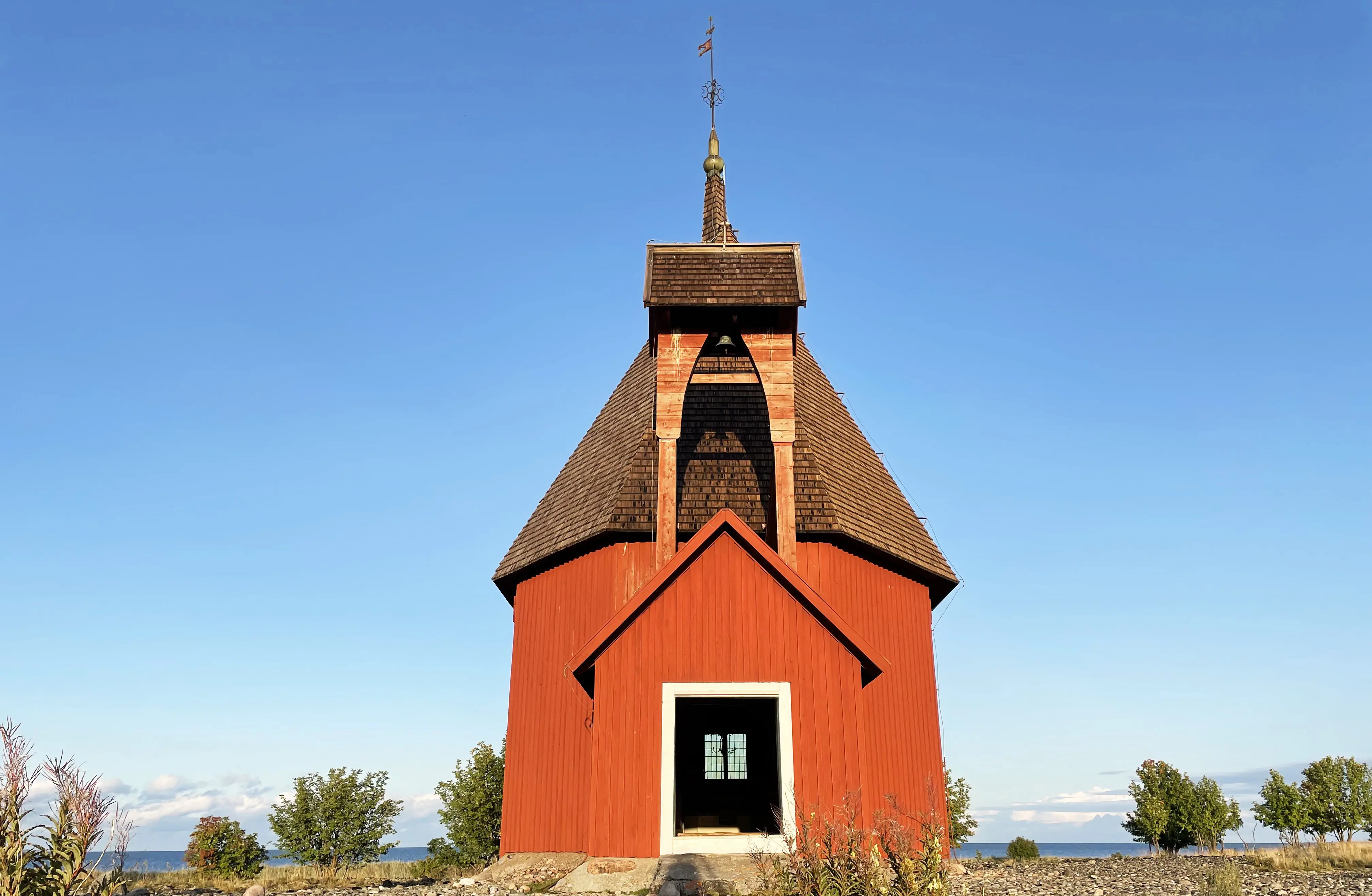 Malörens kapell. En rödmålad träbyggnad med spåntak, omgiven av klappersten och låga buskar. I bakgrunden havet.