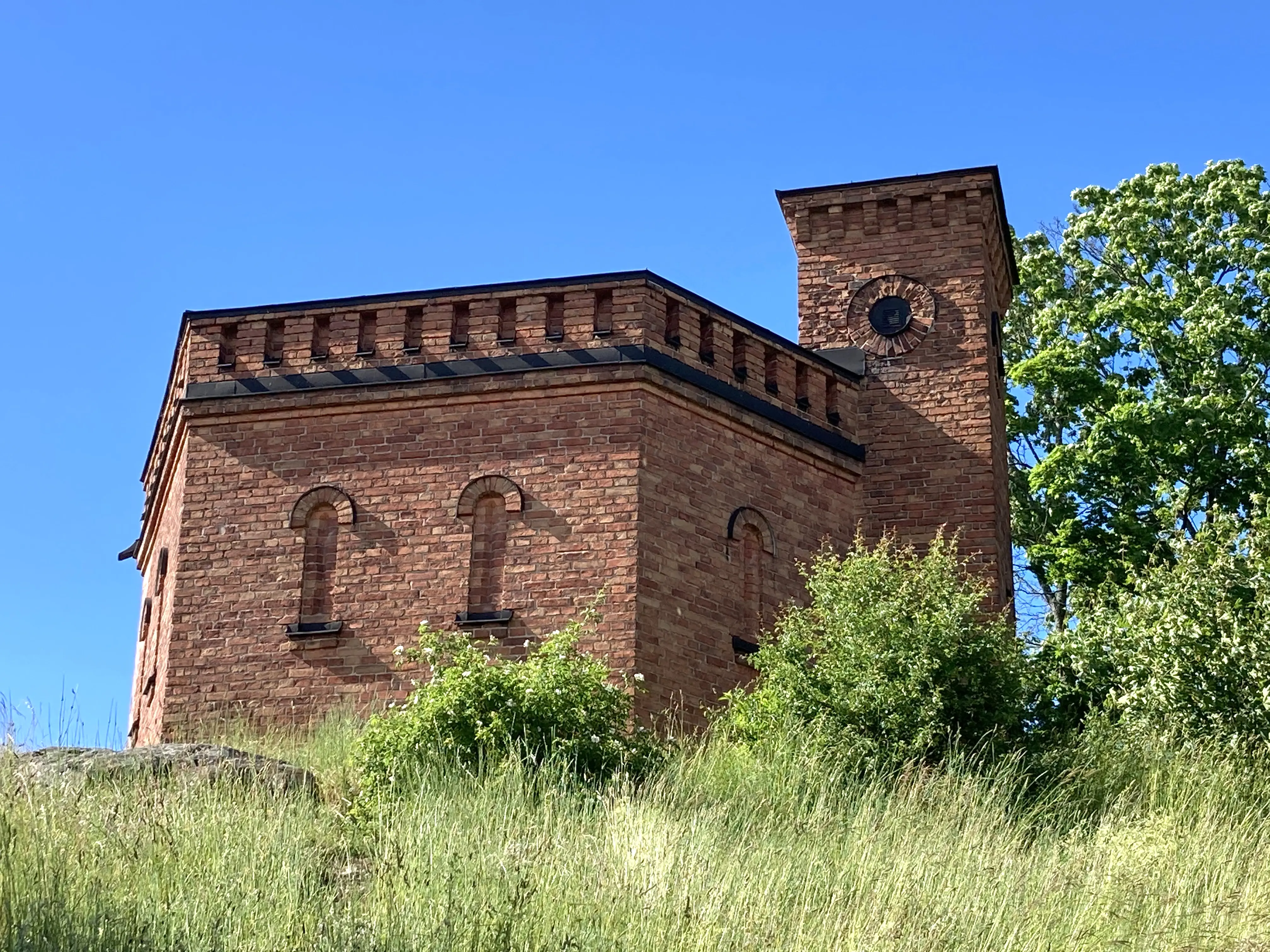 Vattentornet på Skeppsholmen. Sexkantig tegelbyggnad som liknar en medeltida borg med kreneleringar och torn. Sommar och grönska.