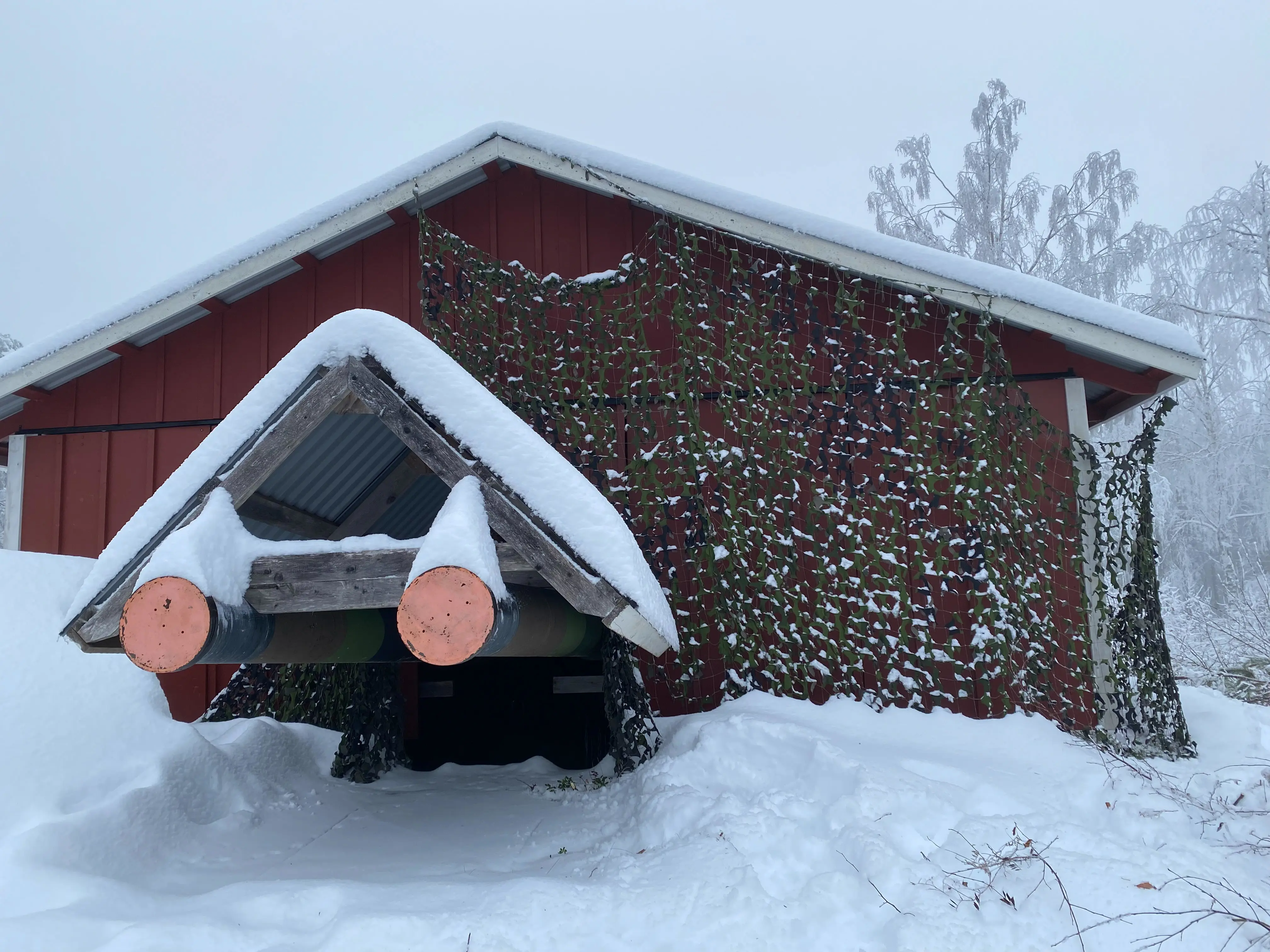 Bilden visar ett rött litet hus med mycket snö. Huset saknar fönster och har ett grönt kamoflagenät fästat på delar av väggen. Rakt fram i bilden sticker ett extra tak ut. Under detta sticker det ut två kanonmynningar pluggade med röda metallskivor.