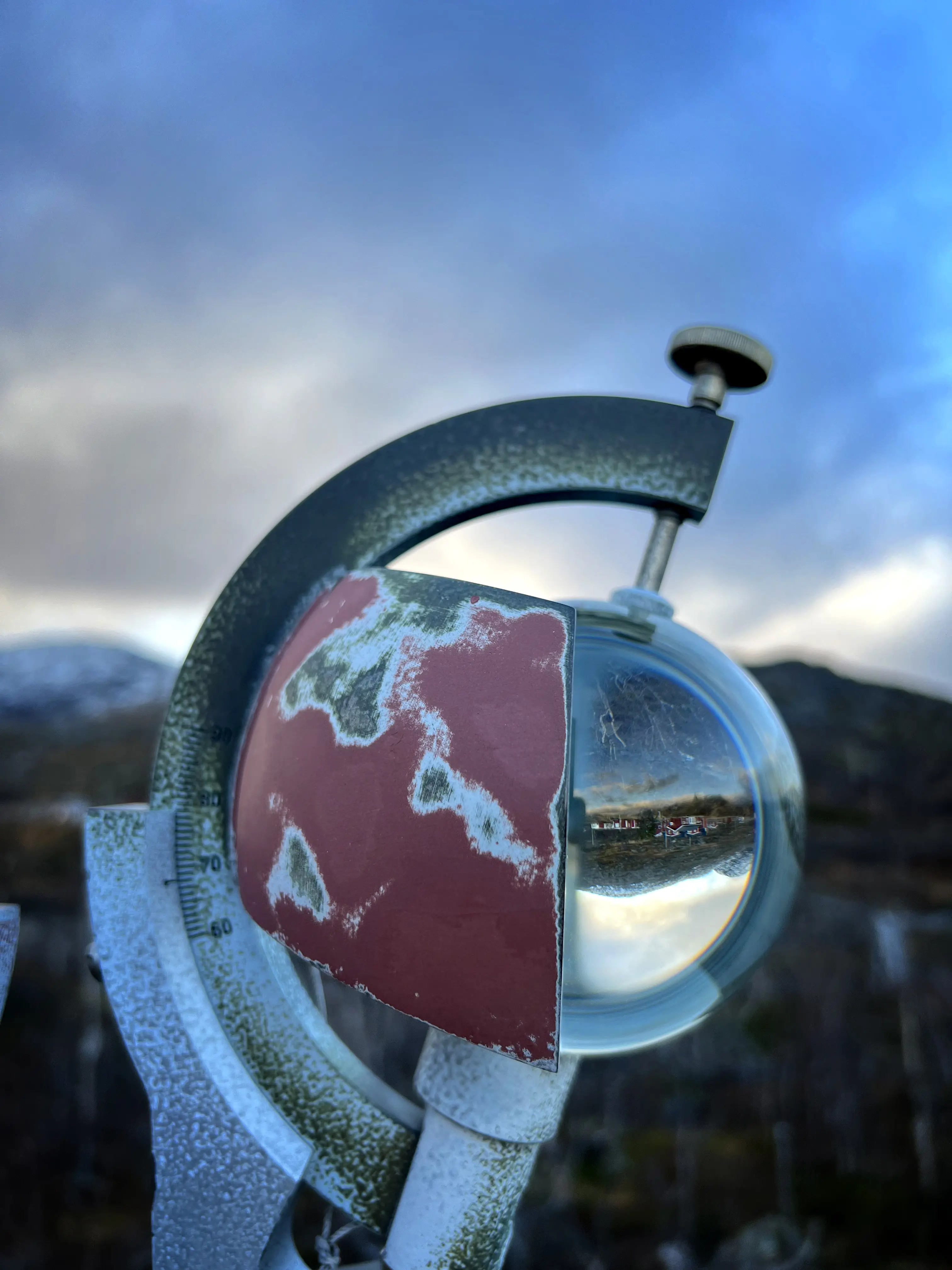 En glaskula sitter i en hållare. I glaset speglas omgivningen med natur och röda hus.