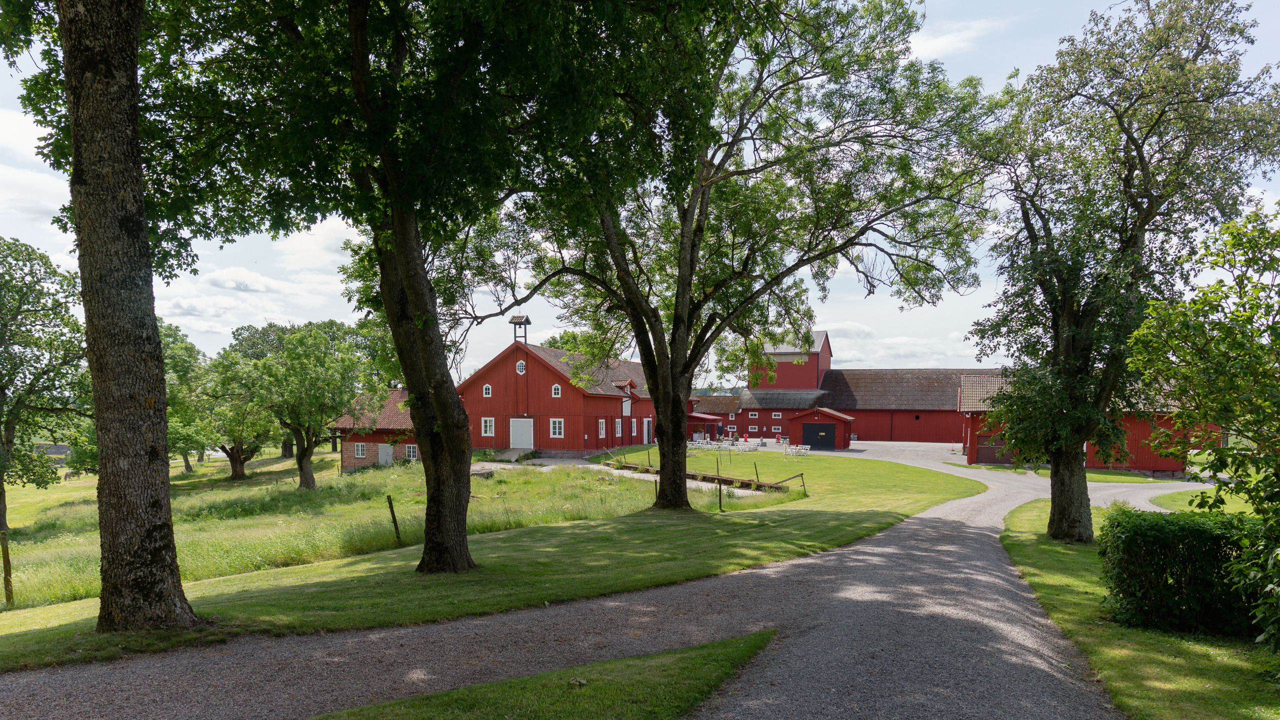 Landsberga. En liten allé med grusgång leder mot en samling röda ekonomibyggnader av olika storlekar och ålder.