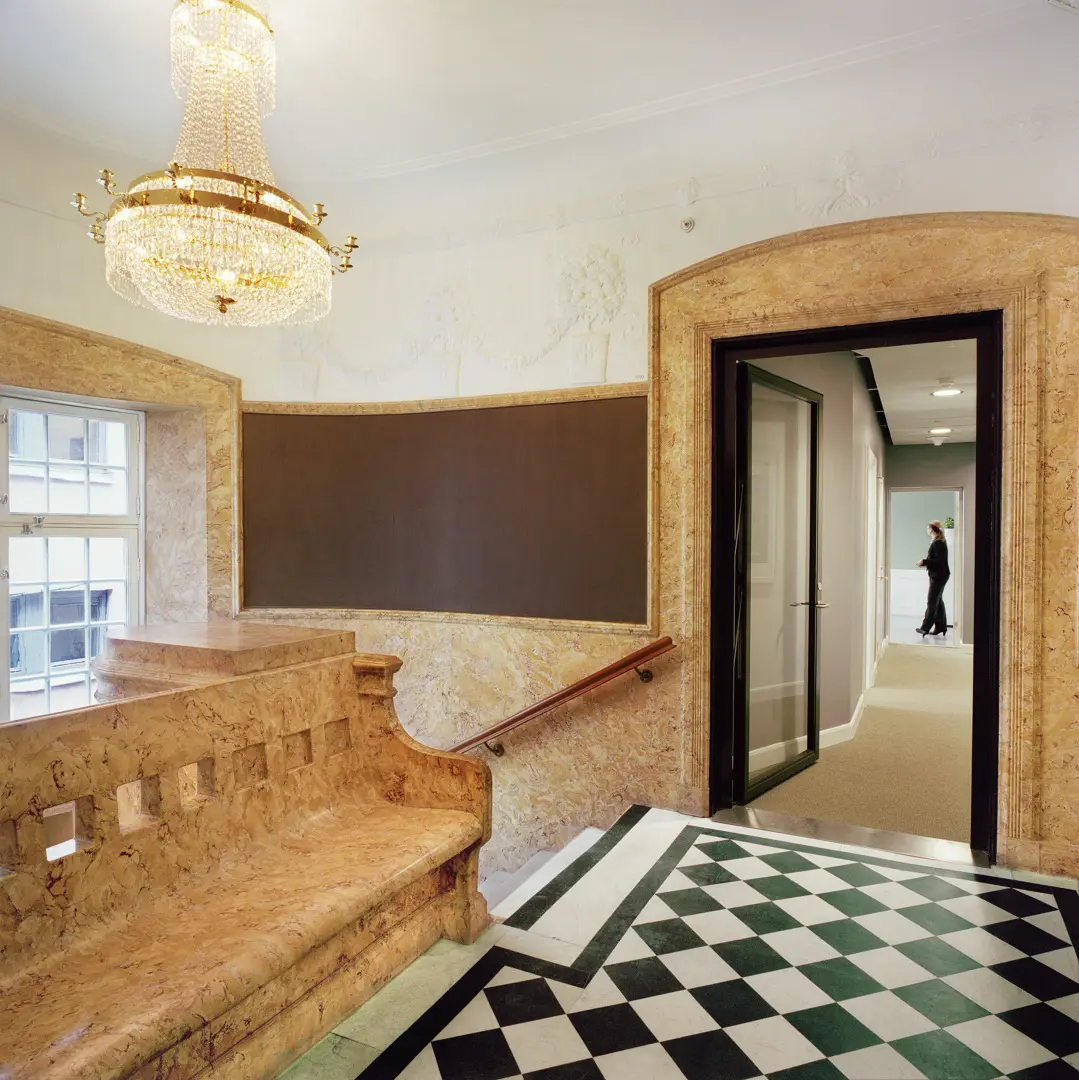 Trapphus med kristallkrona, svartvitrutigt golv. Bänk och väggar är av ett rödaktigt marmorerat stenmaterial.