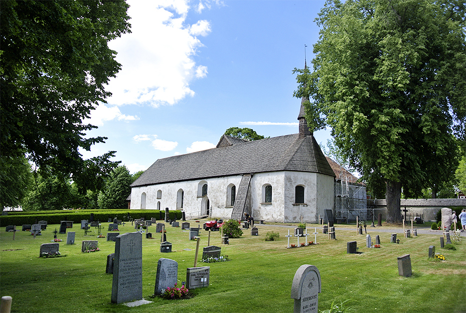 En vit avlång kyrka med ett ljust grått tak och ett litet torn med spira. Framför kyrkan syns en gräsmatta med gravstenar och kors..
