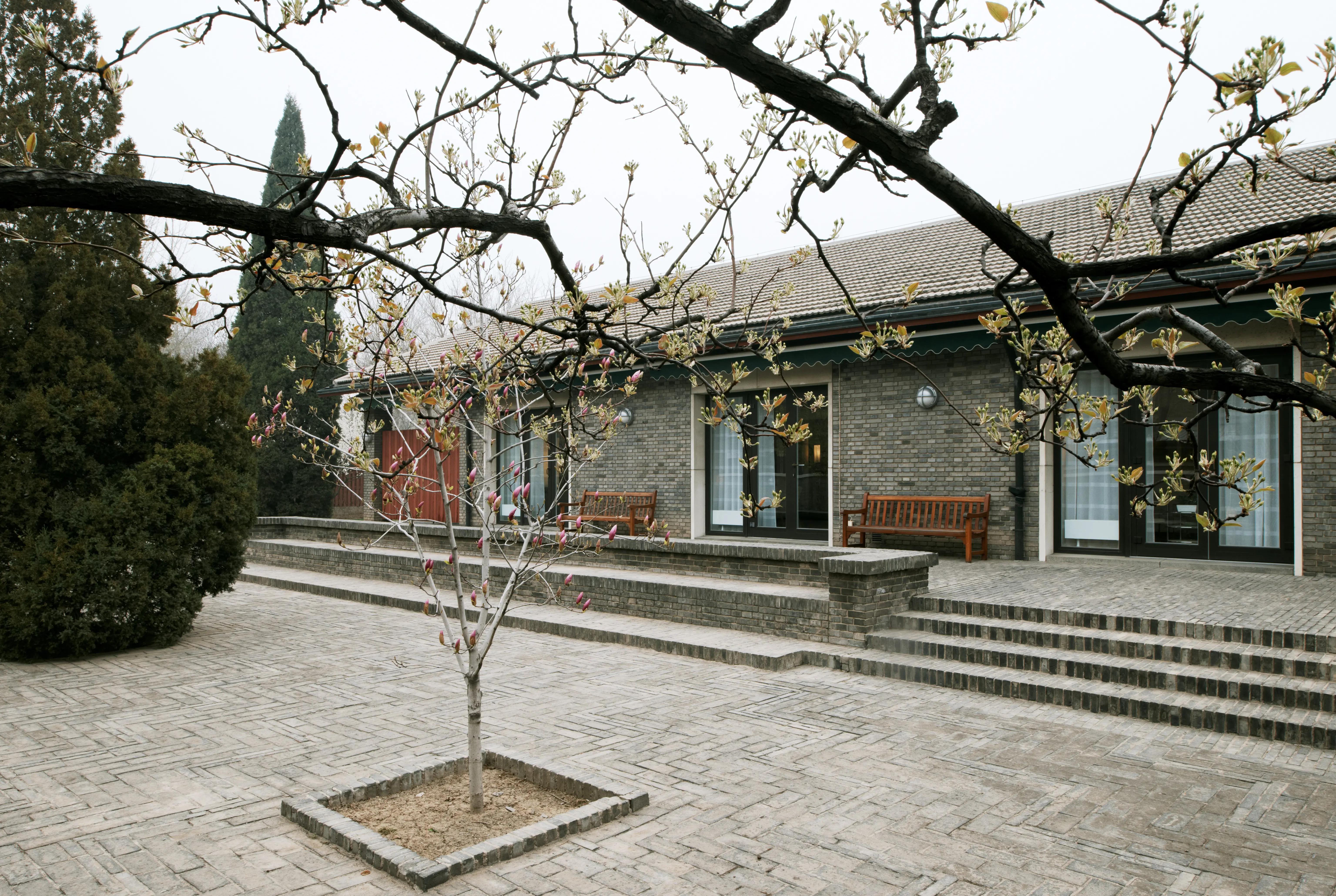 Ambassaden i Peking. Fasad mot en stenlagd gård med enstaka träd. Vår.