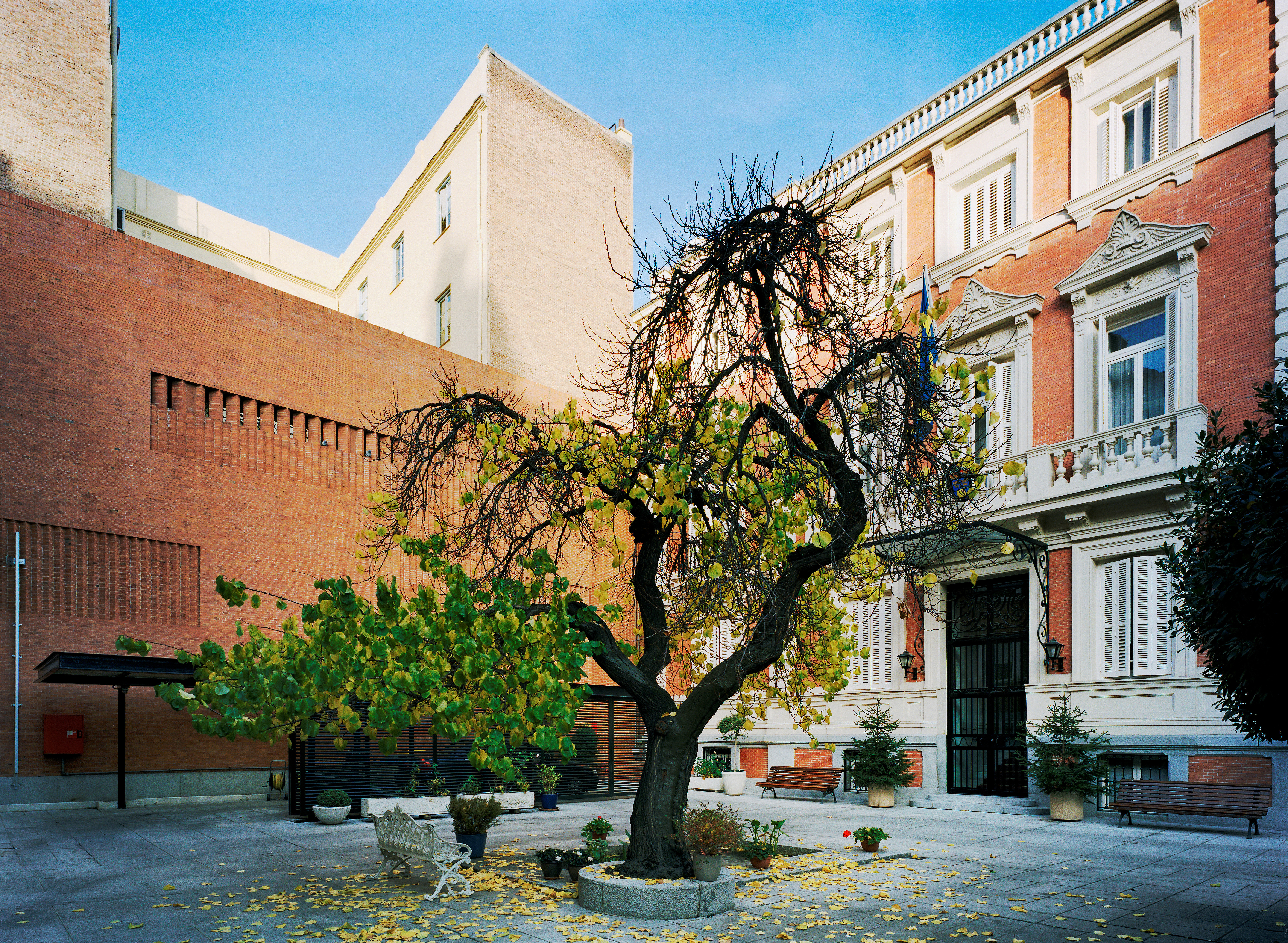 Ambassaden i Madrid. Stenlagd innergård med ett träd och en liten fontän. En hög, modernistisk tegelmur möter den äldre, utsmyckade byggnaden.