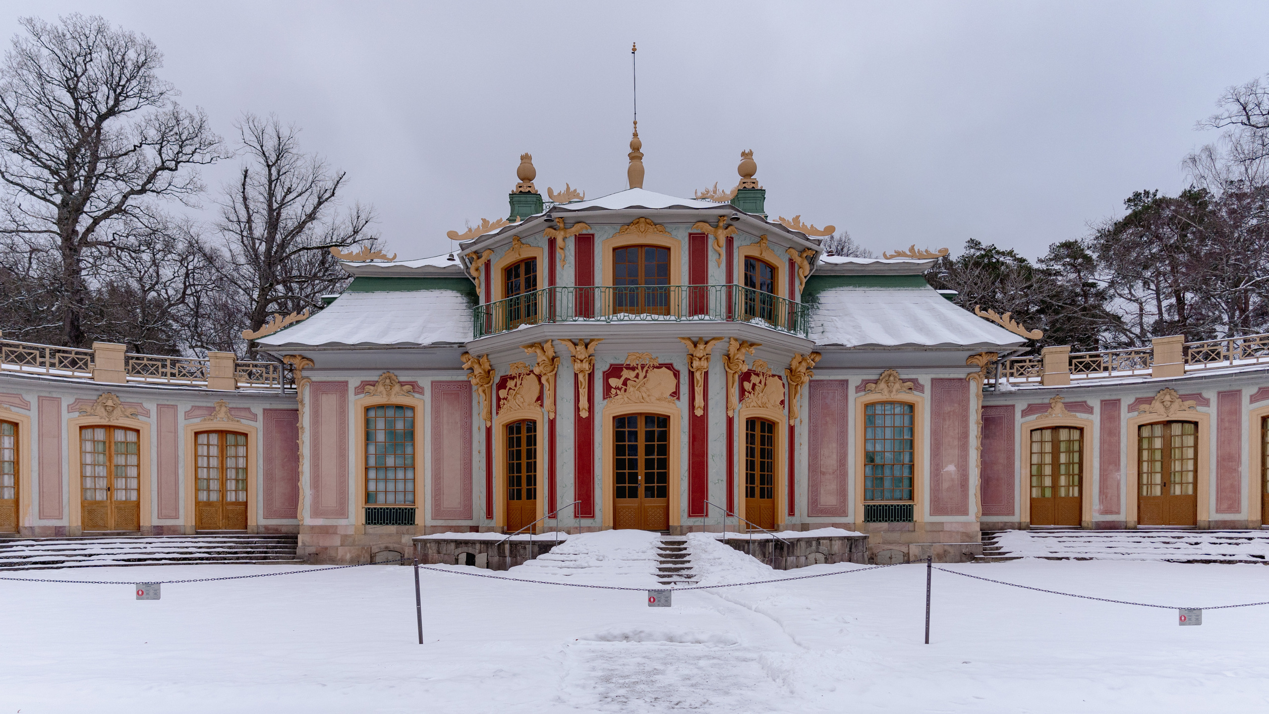 Kina slott, Drottningholm. Litet rosa slott med grönt, vågigt plåt-tak. Fasaden har skulpterad, gul dekor. Vinter och snö.