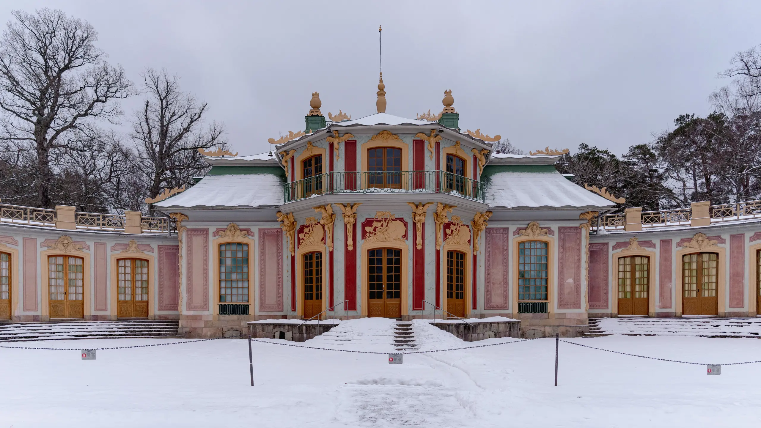 Kina slott, Drottningholm. Litet rosa slott med grönt, vågigt plåt-tak. Fasaden har skulpterad, gul dekor. Vinter och snö.