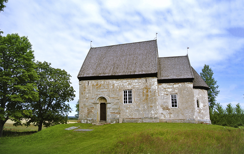 En stenkyrka med brunaktigt tak. Kyrkan står på en liten upphöjd gräskulle och omges av grönska och blå himmel.