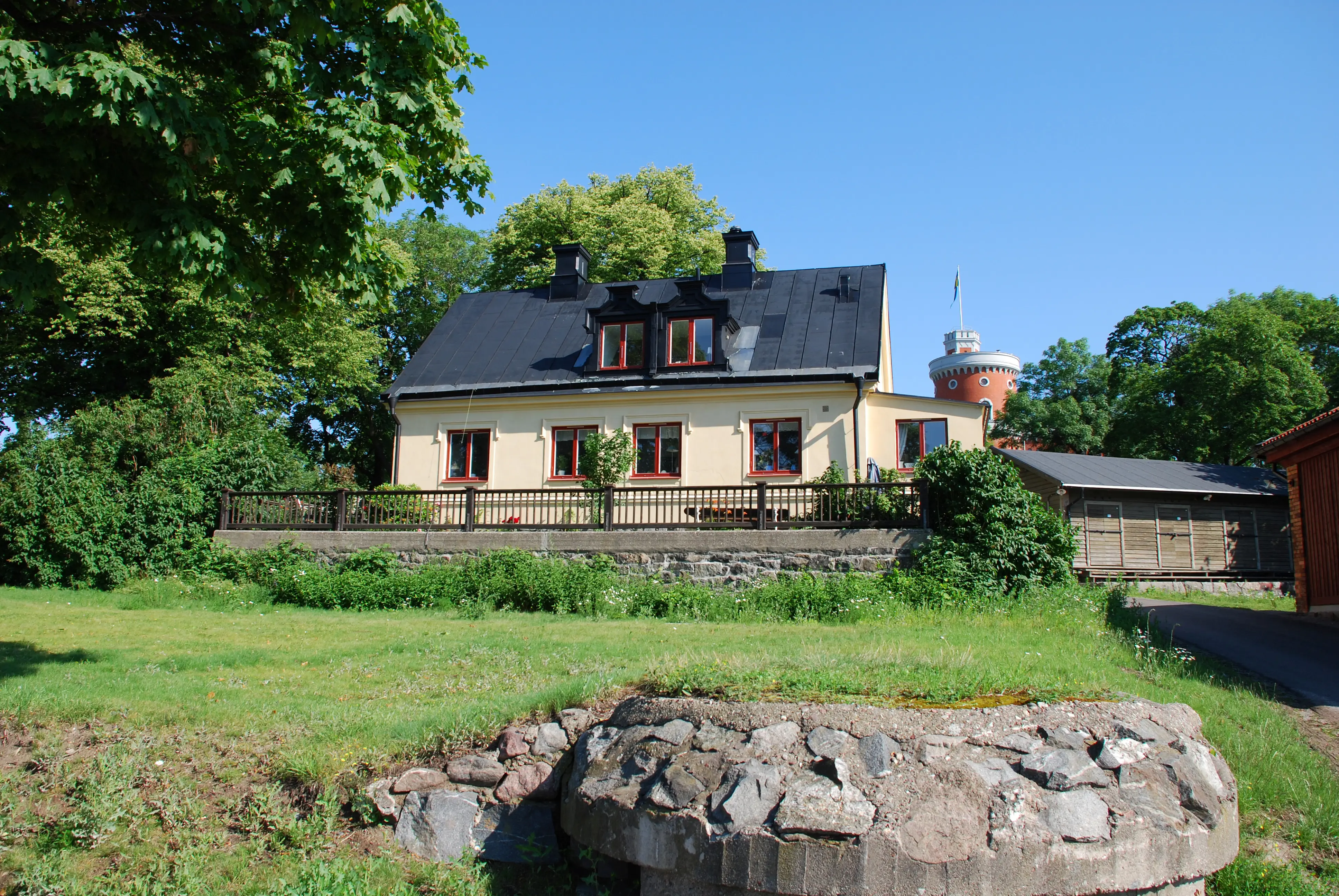 Flaggkonstapel-bostället på Kastellholmen. Gulputsad villa med lummig liten trädgård. Det svarta plåt-taket har två tak-kupor.