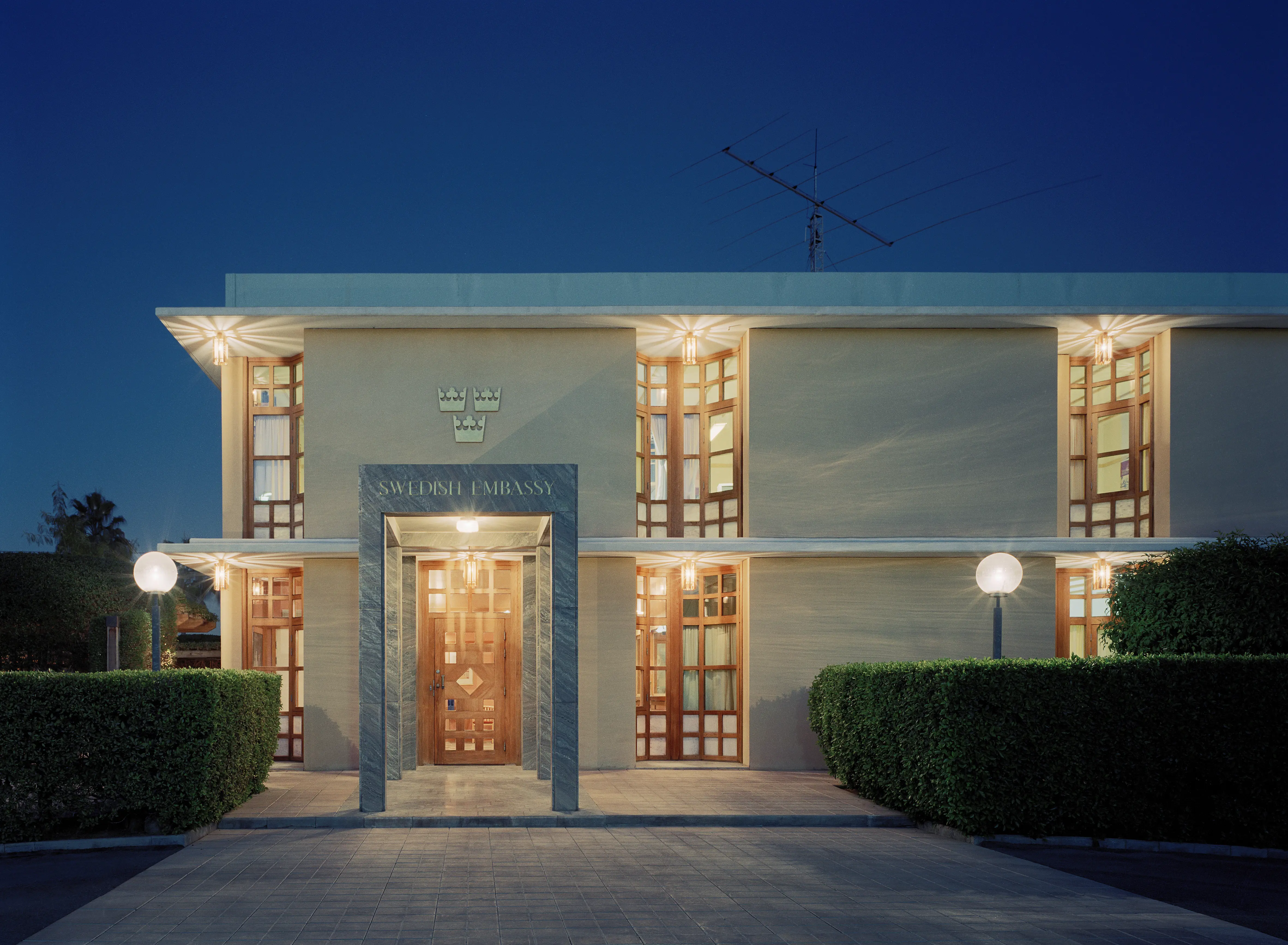 Ambassaden i Riyadh. En vitputsad tvåvåningsbyggnad med rundade, höga fönsternischer. Entrén ramas in av en stenportik. Kväll.