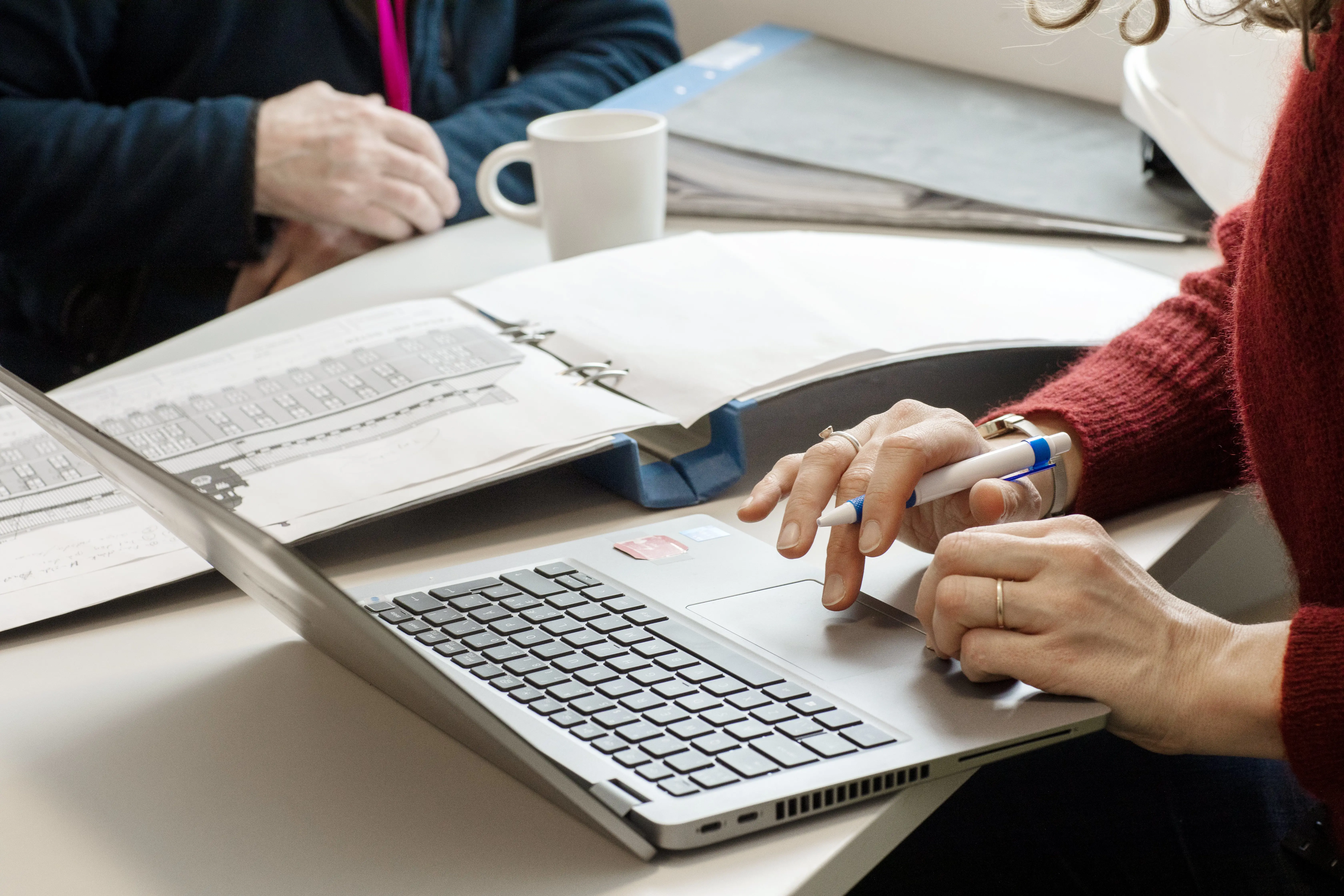 Två personer sitter vid ett bord med laptop och uppslagna pärmar.
