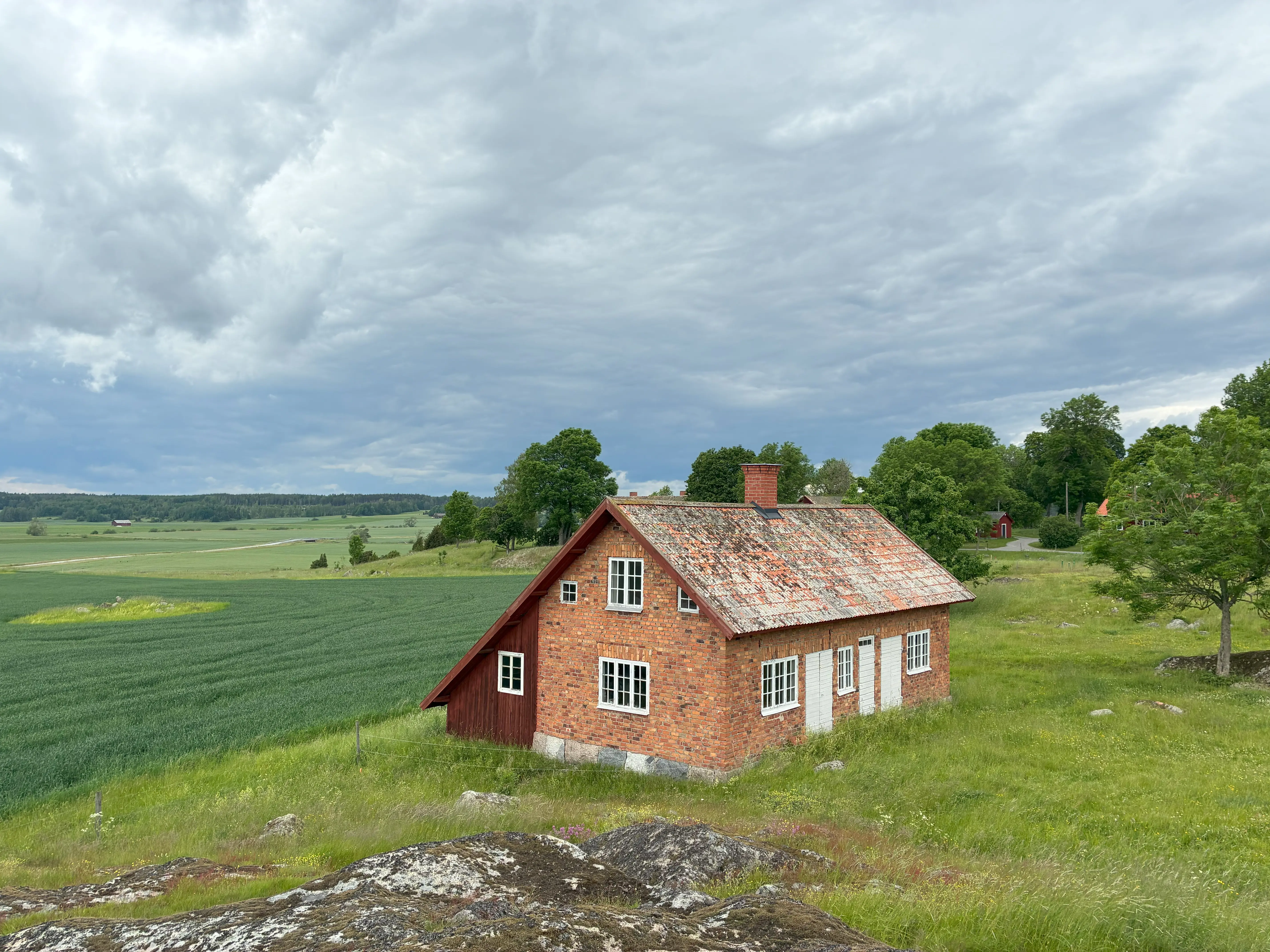 Landsberga. Tegelbyggnad ute på ett uppvuxet betes-fält. I bakgrunden brer jordbruks-landskapet ut sig.
