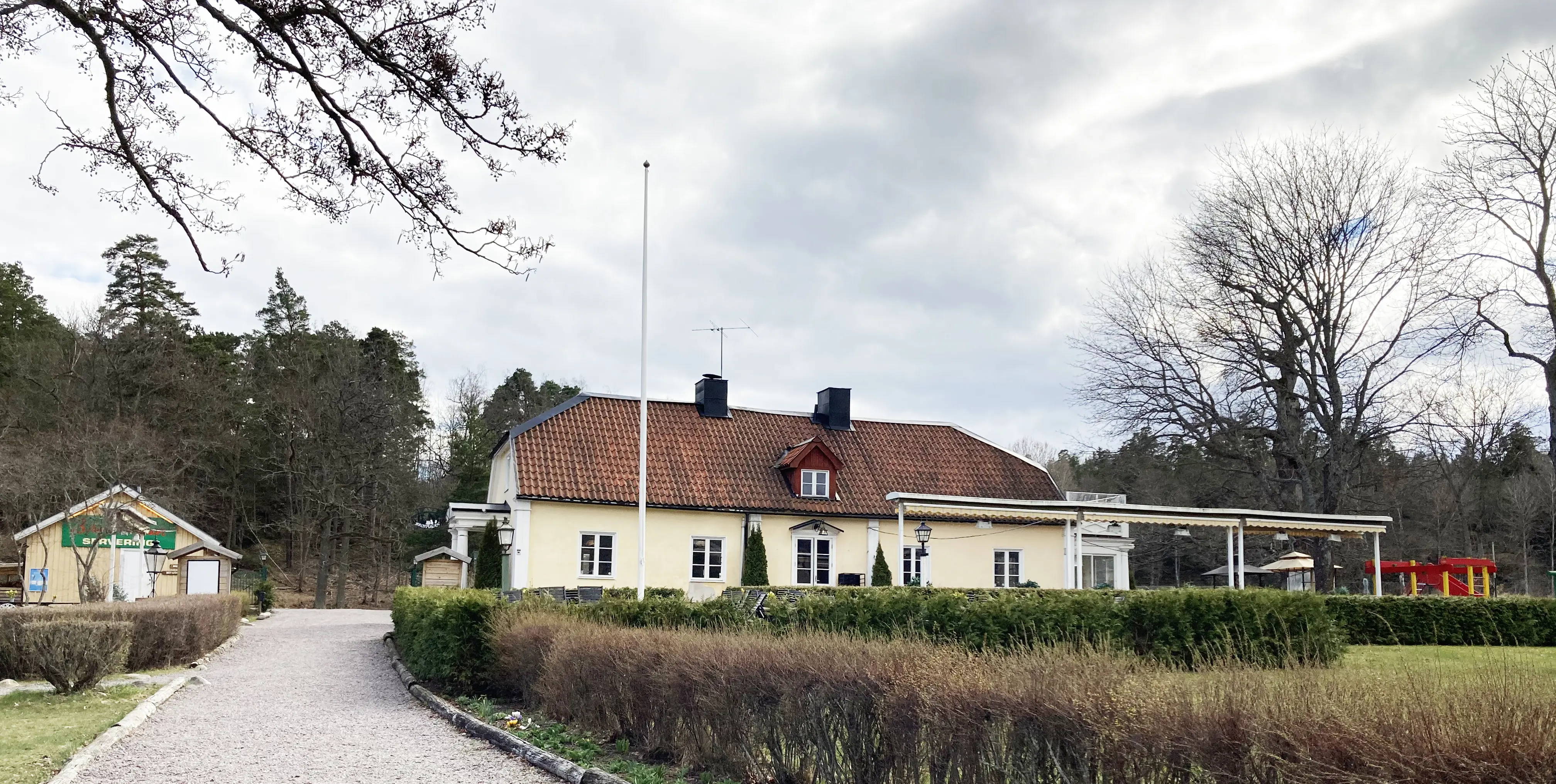 Brostugan på Kärsön. En gulputsad tvåvåningsbyggnad med brutet tegeltak, omgivet av en trädgårds-servering. Tidig vår.