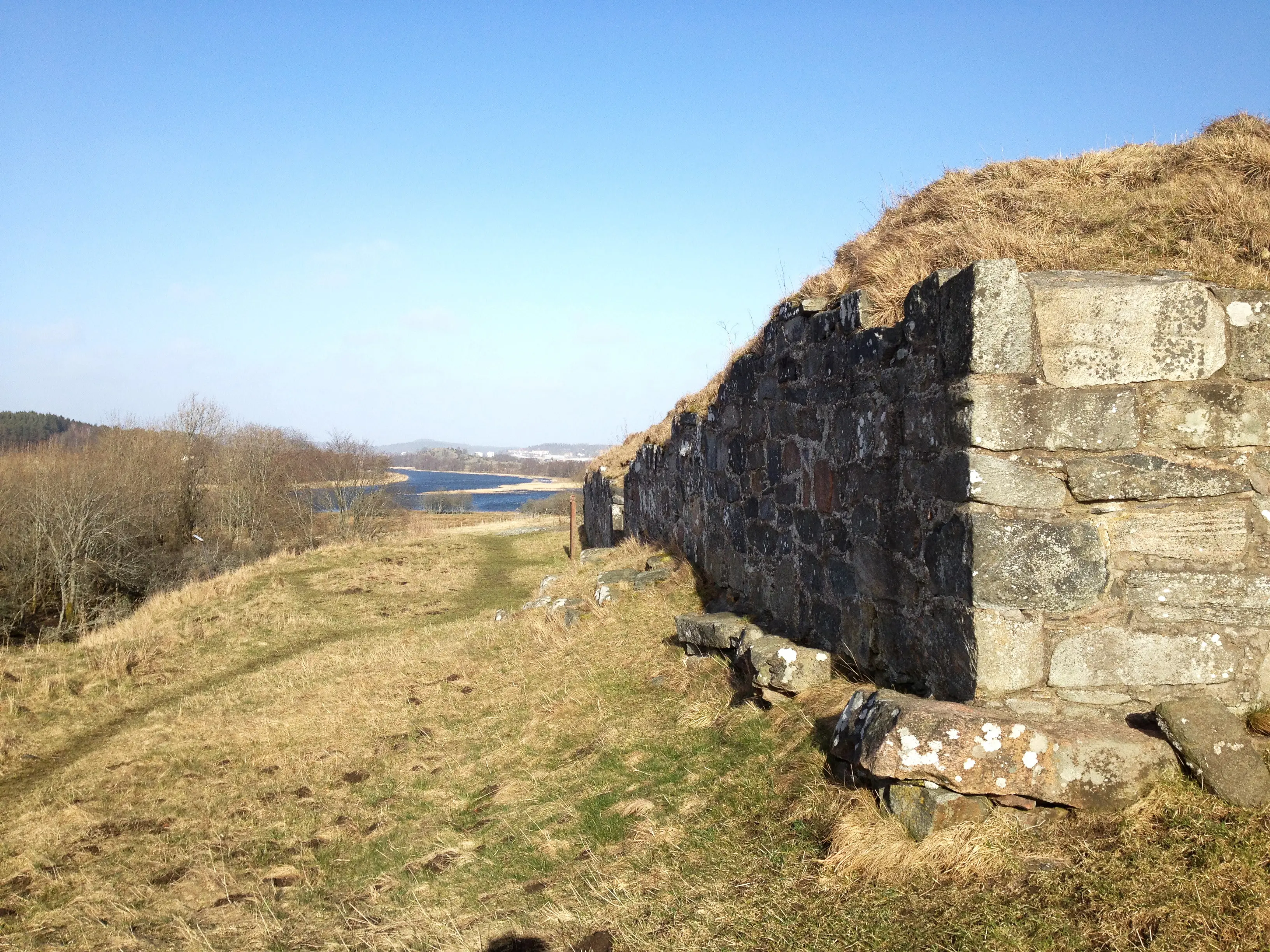 Ragnhildsholmens borgruin. Gräsbevuxna rester av stenmurar. Vy mot Nordre älv.