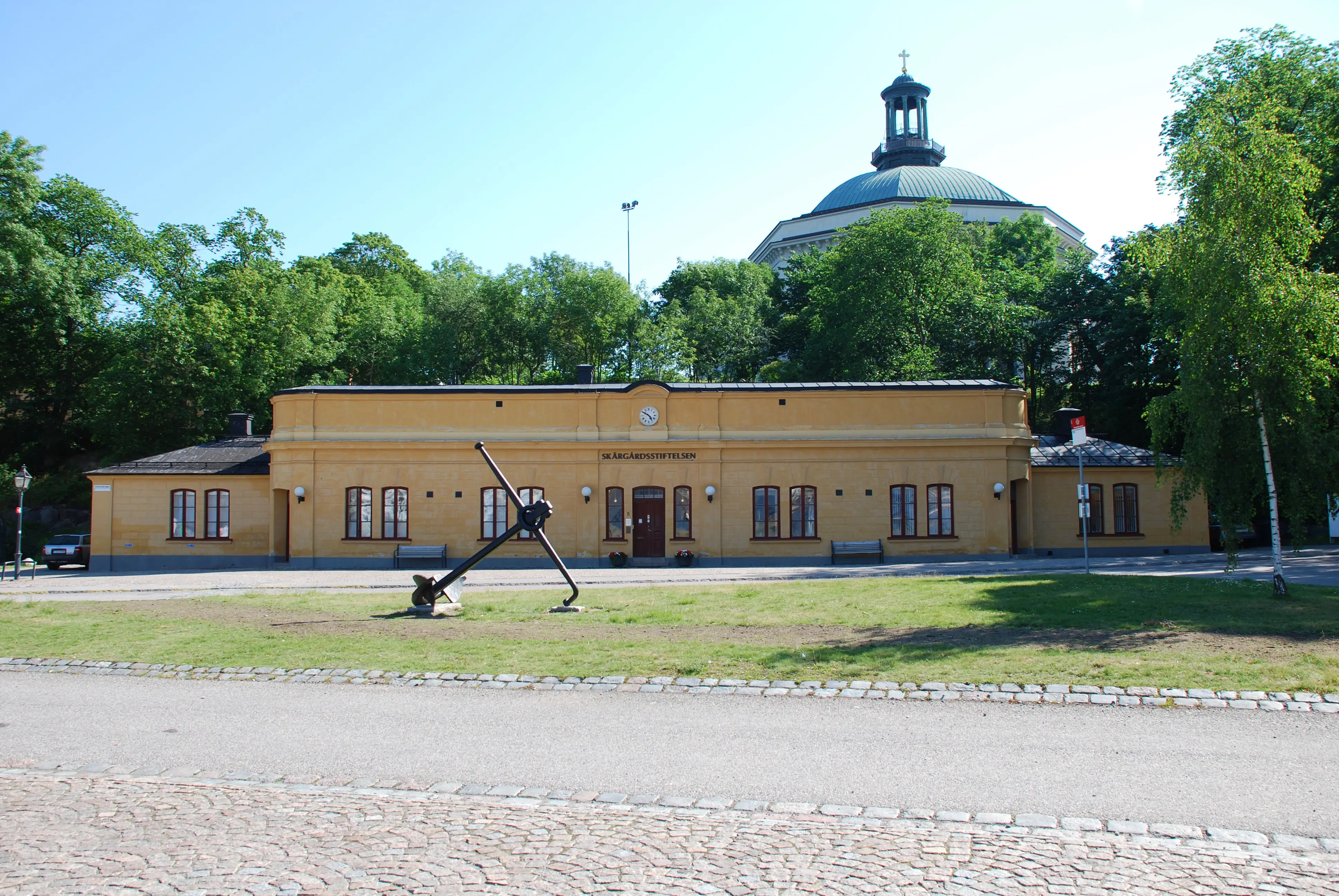 Högvakten på Skeppsholmen. Låg, gulputsad byggnad i enkel nybarock-stil. I bakgrunden syns Skeppsholmskyrkan. Sommar och grönska.