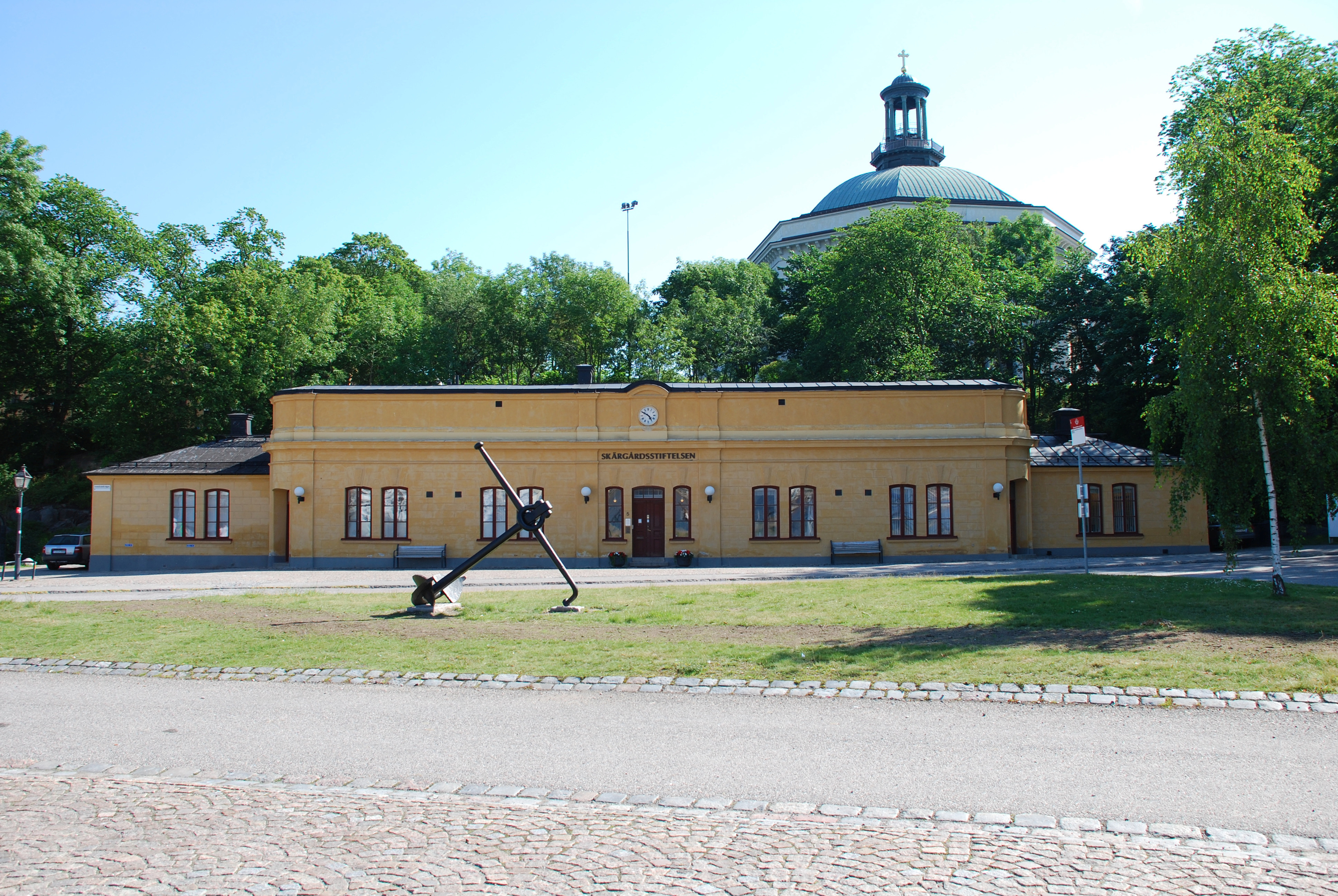 Högvakten på Skeppsholmen. Låg, gulputsad byggnad i enkel nybarock-stil. I bakgrunden syns Skeppsholmskyrkan. Sommar och grönska.