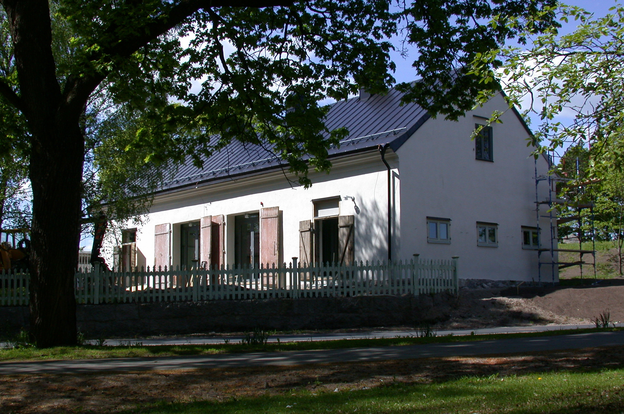 Landshövdingens före detta stall. Ljusgrå byggnad med svart plåt-tak. Fönstren har enkla träluckor. Sommar och grönska.
