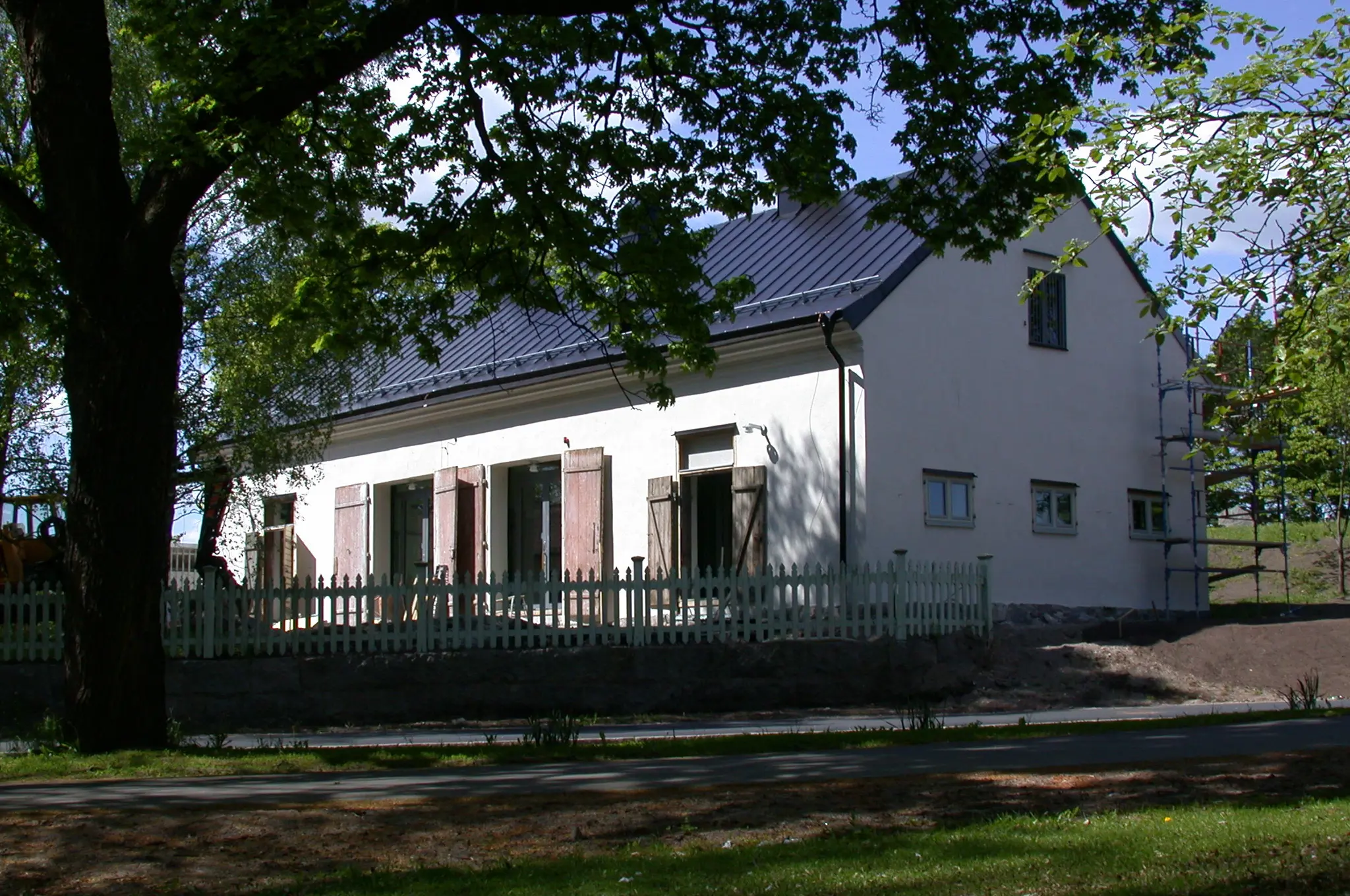 Landshövdingens före detta stall. Ljusgrå byggnad med svart plåt-tak. Fönstren har enkla träluckor. Sommar och grönska.