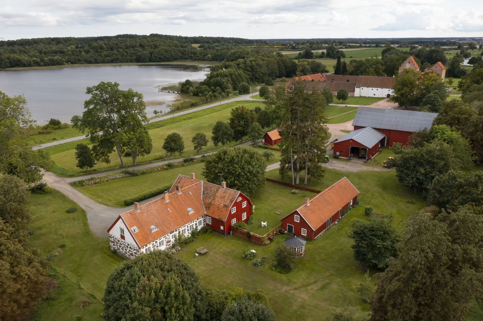Flygbild av kungsgården. En delvis vitputsad stenbyggnad och några rödmålade träbyggnader med en sjö i bakgrunden.