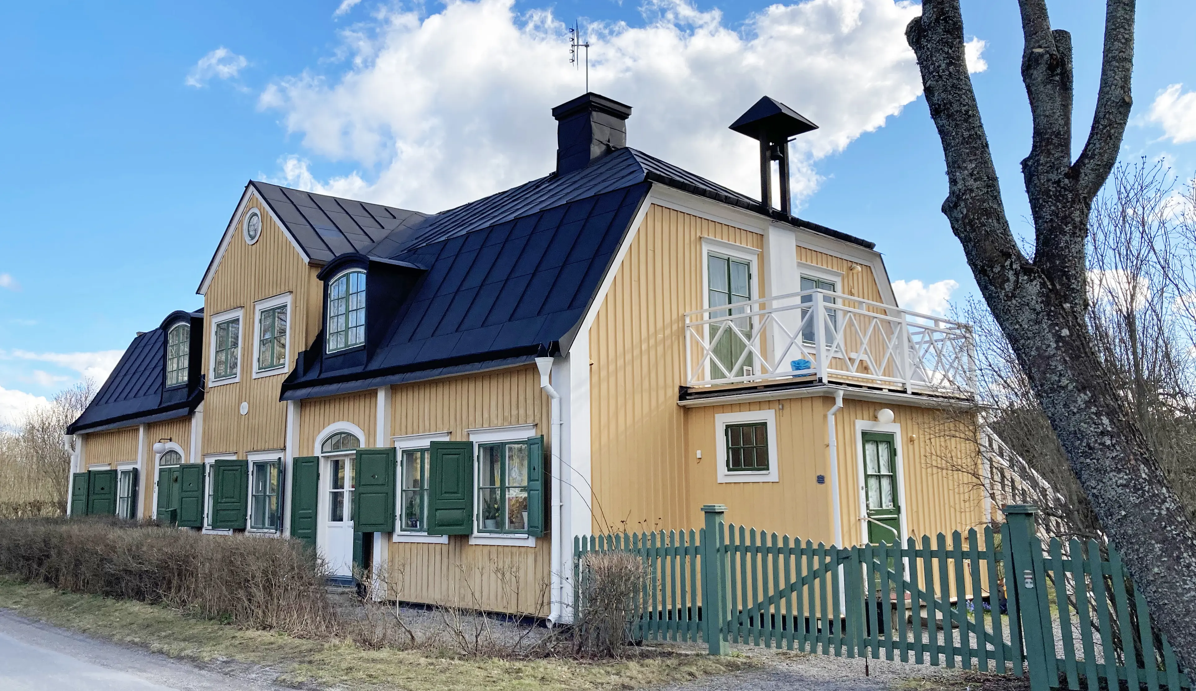 Kanton 2. Gult parhus med mittgavel och gröna fönsterluckor. Det svarta plåt-taket har två tak-kupor. Gaveln har en utbyggnad med balkong.