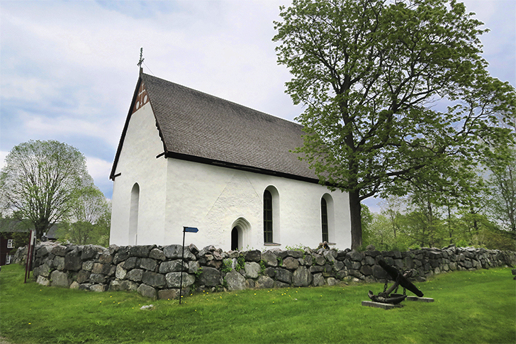 En fyrkantig liten stenkyrka med vitpustad fasad och brunt sluttande tak. Kyrkan omges av en stenmur.