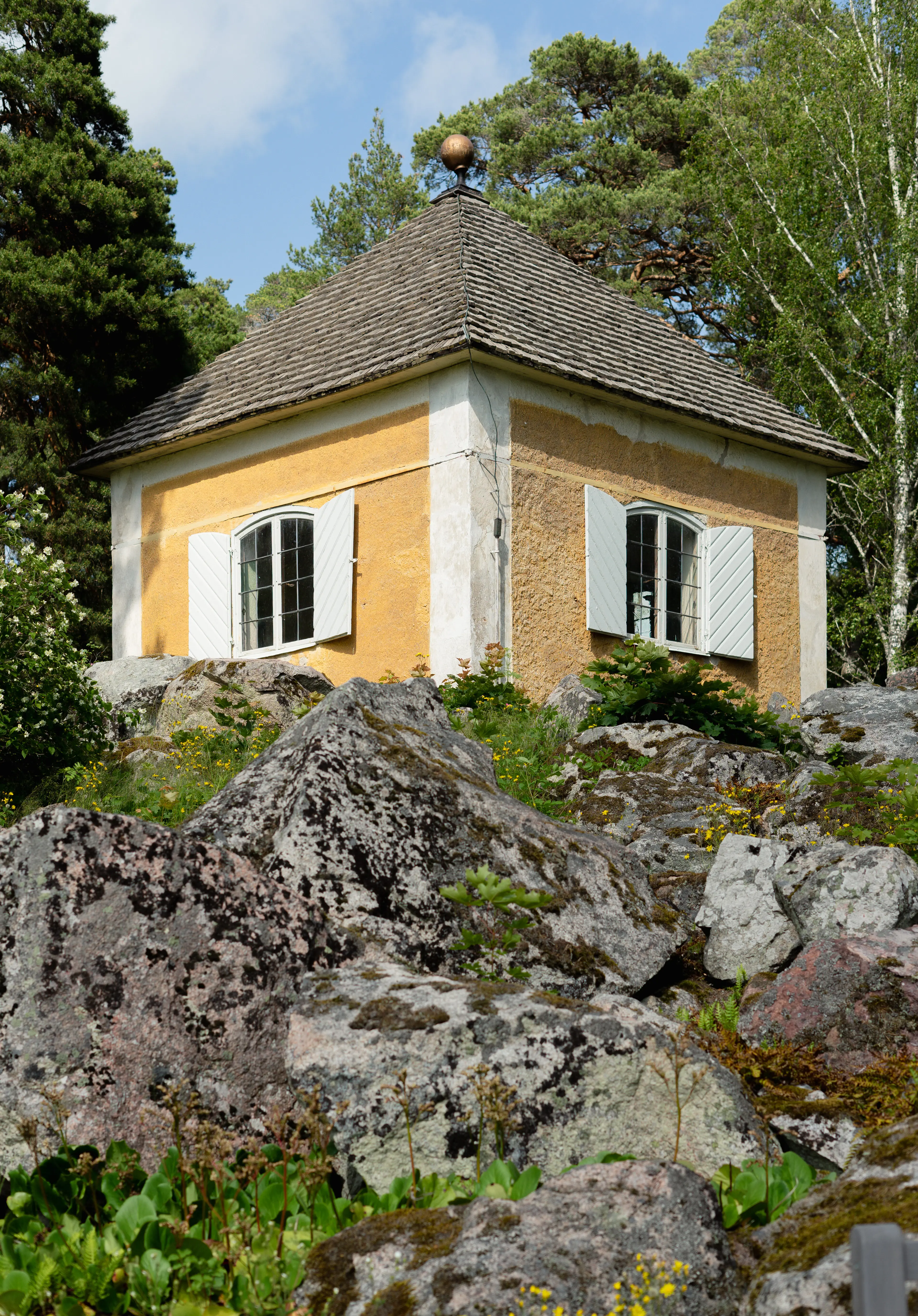 Museibyggnaden på Linnés Hammarby. En gul paviljong med pyramid-format spån-tak uppe på en stenig höjd.