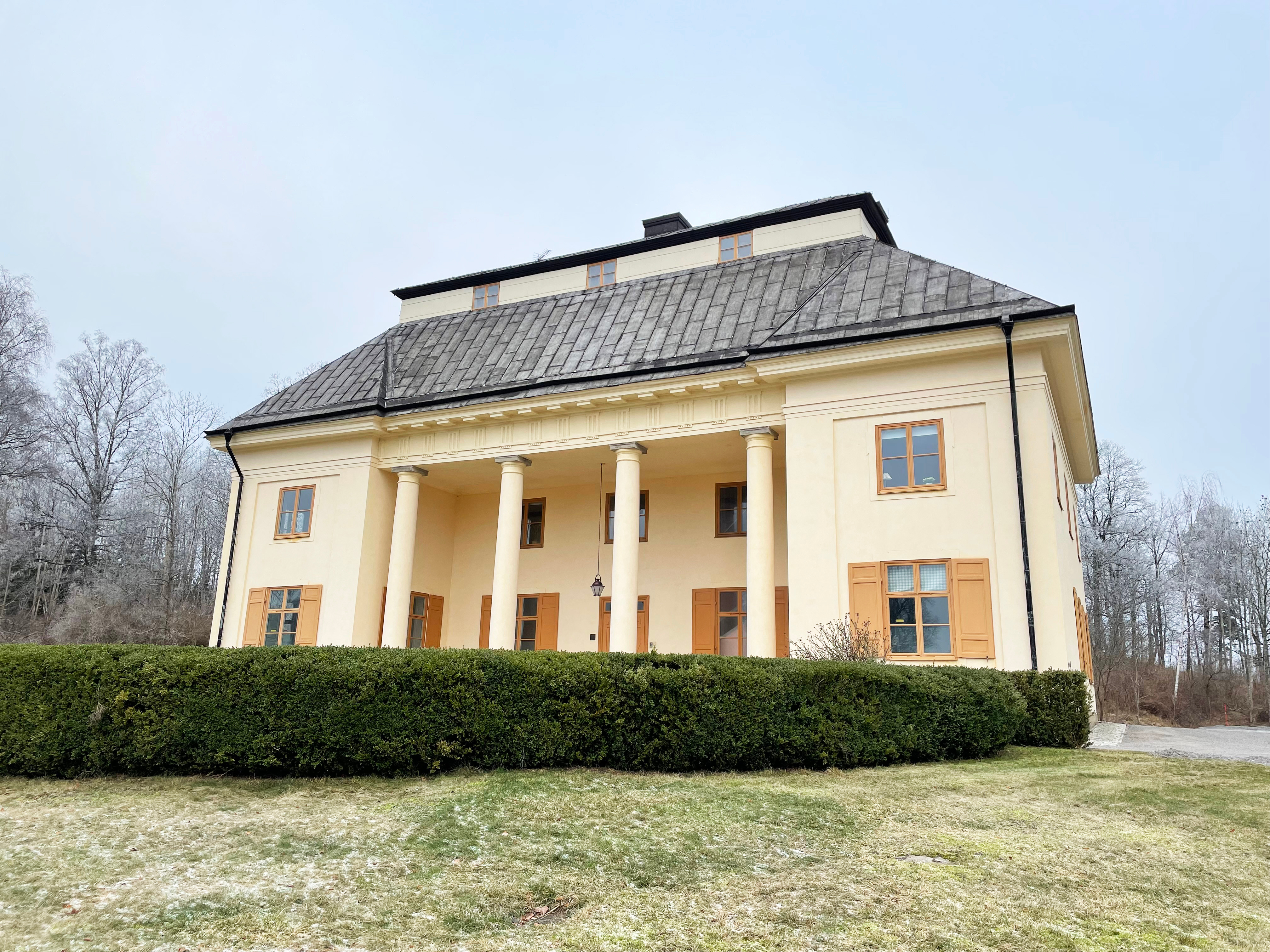 Hemmet. En ljusgul tvåvångsbyggnad med säteritak och kolonnad. En formklippt häck avgränsar en del av trädgården framför huset.