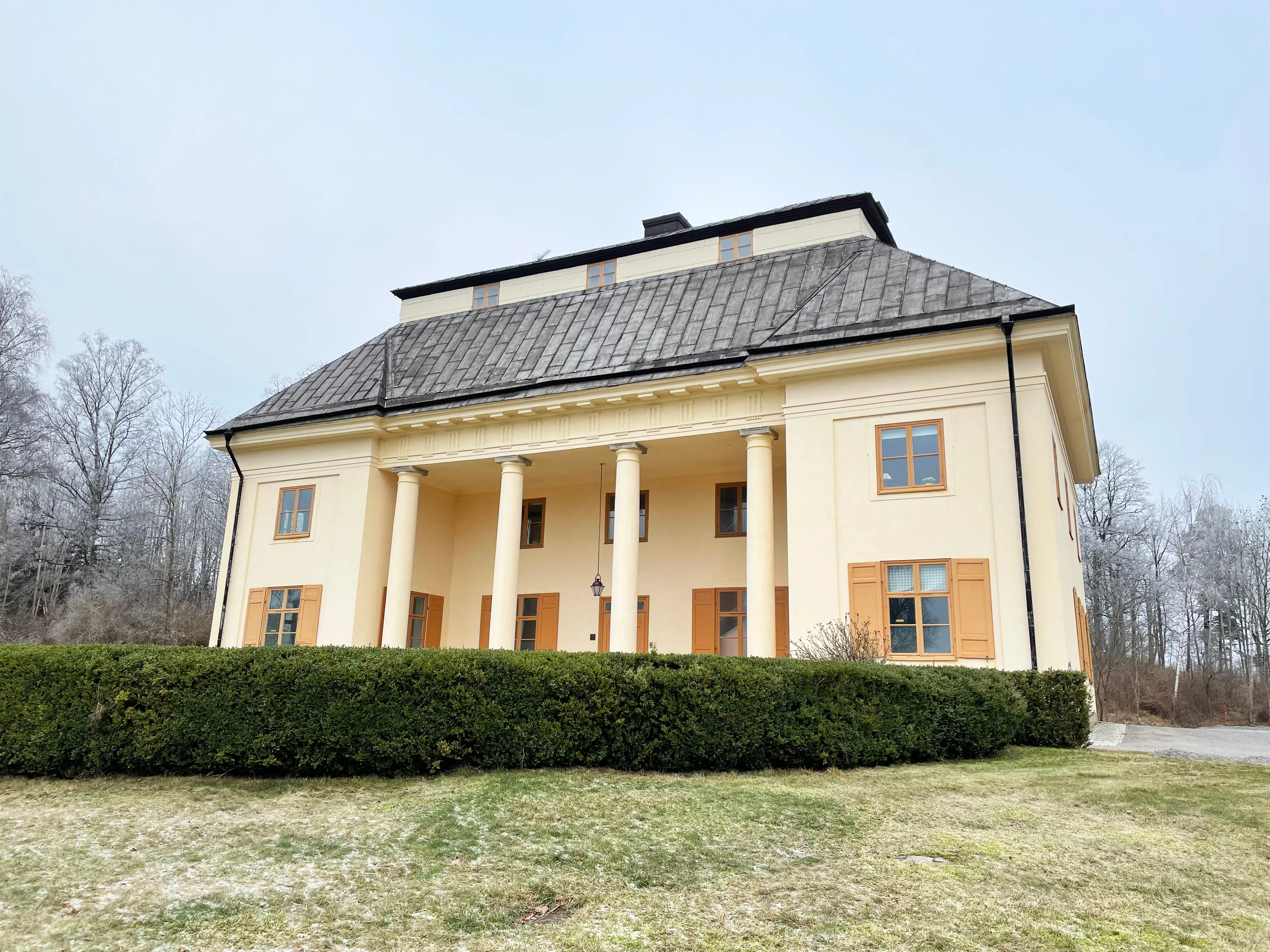 Hemmet. En ljusgul tvåvångsbyggnad med säteritak och kolonnad. En formklippt häck avgränsar en del av trädgården framför huset.