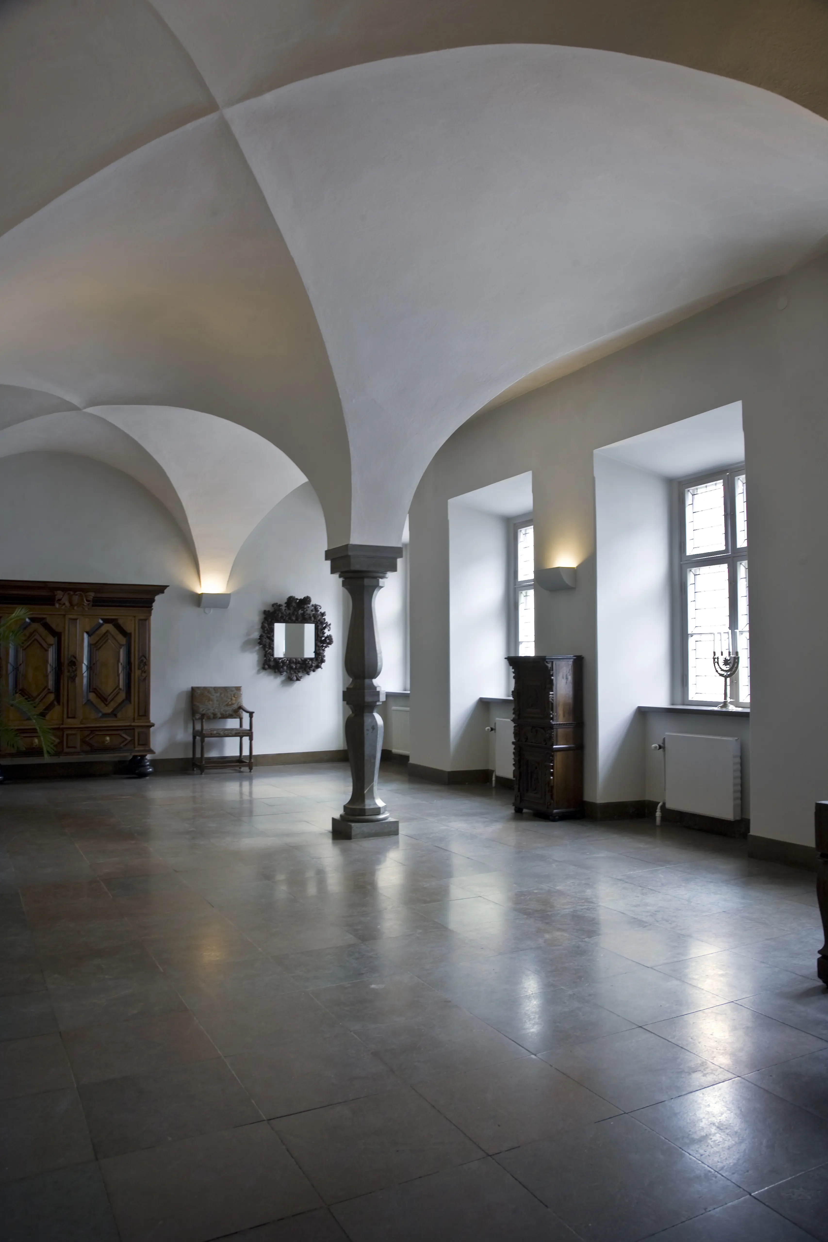 Hall i Kalmar residens. Golvet är av mörk sten. En formhuggen stenpelare bär upp breda, vitputsade valv.