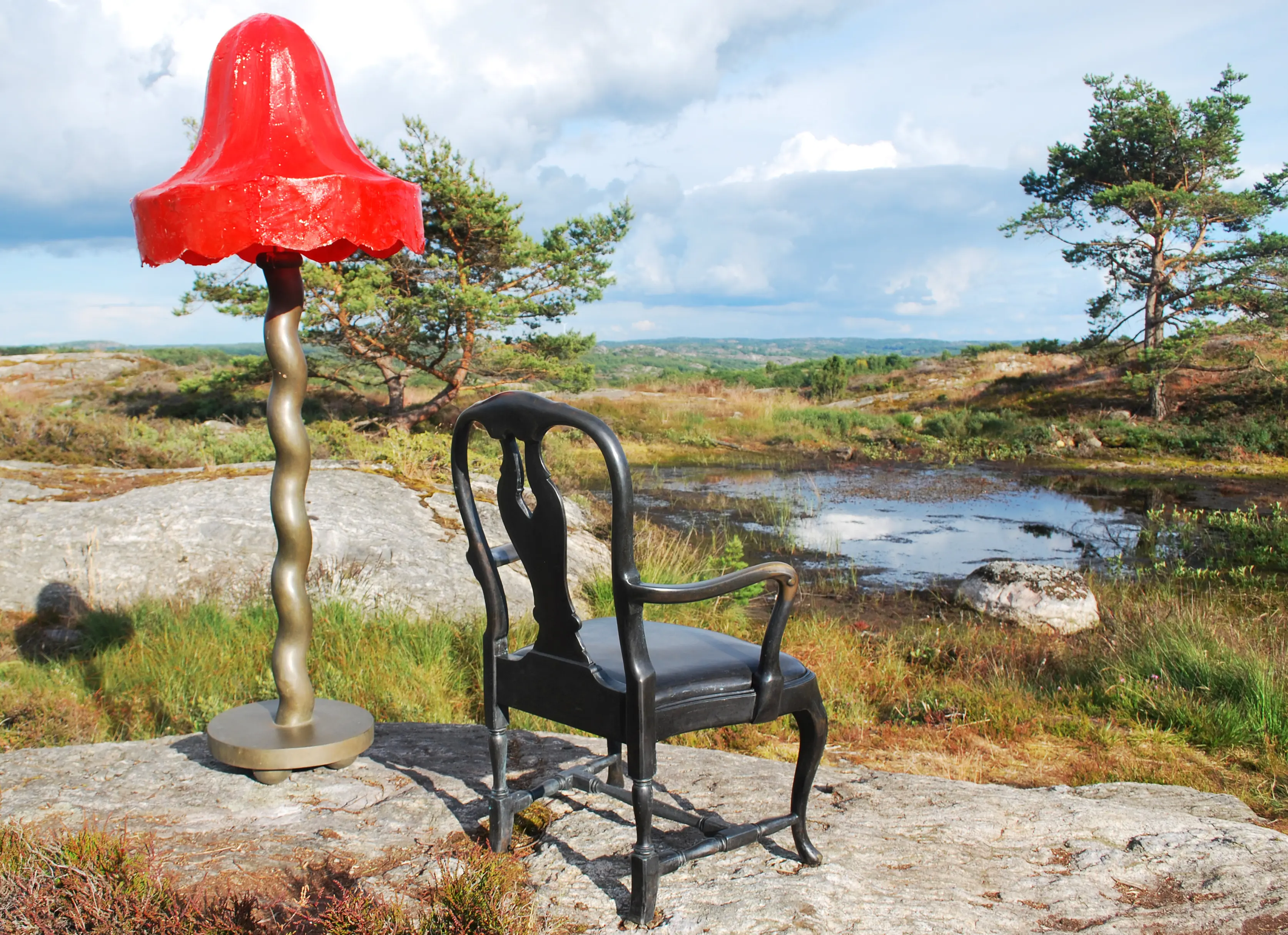 Pilane. På en stenig höjd med en vattensamling står skulpturer i form av en golvlampa med röd skärm och en svart karmstol.