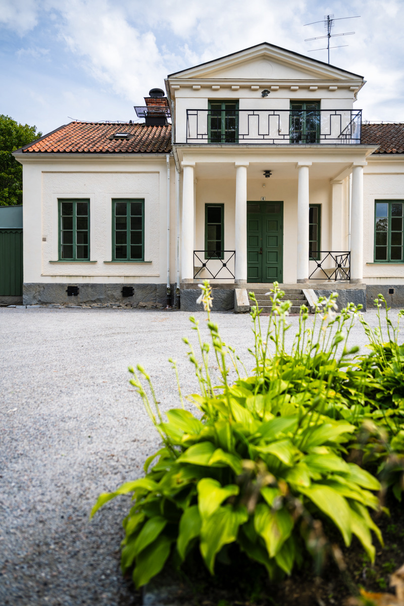 Villa i två våningar med vitputsad fasad och mörkgröna dörrar och fönsterstommar. I förgrunden syns en rabatt på en grusplan.