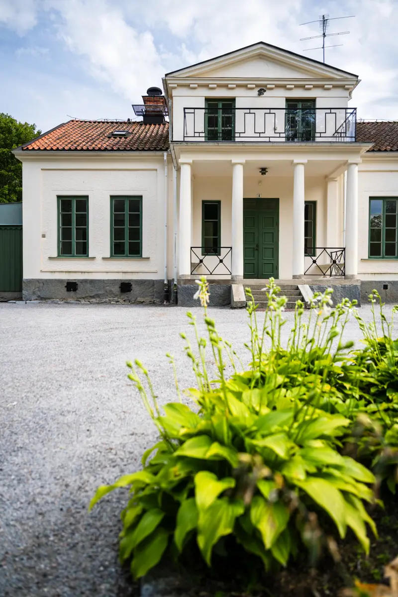 Villa i två våningar med vitputsad fasad och mörkgröna dörrar och fönsterstommar. I förgrunden syns en rabatt på en grusplan.