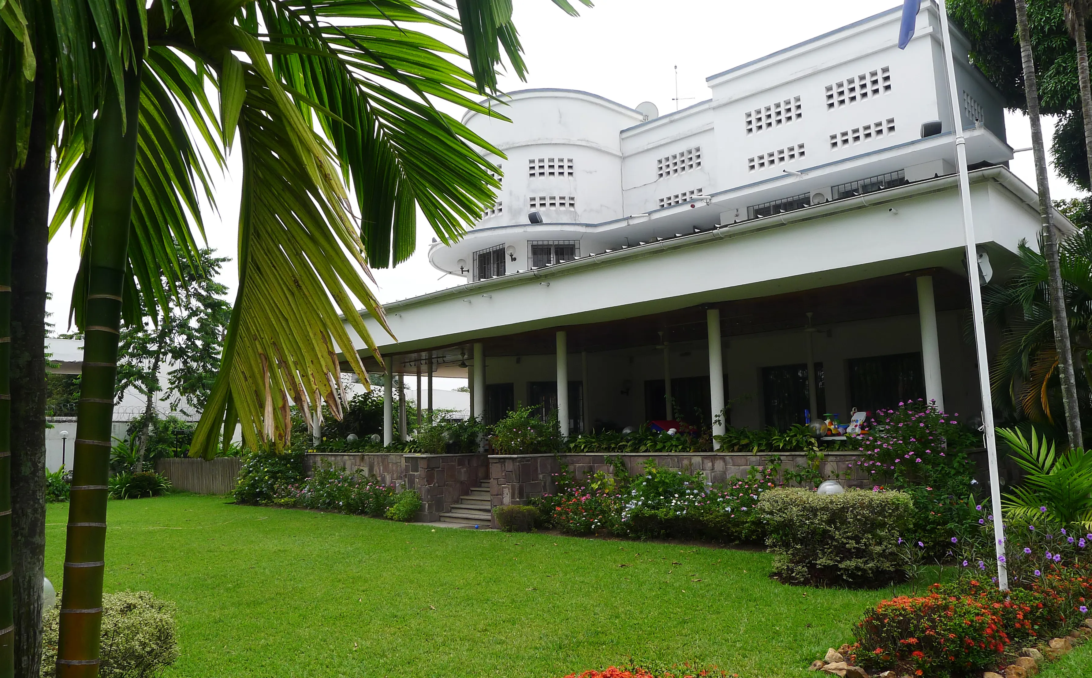 Chefsbostaden i Kinshasa. En vit byggnad i två våningar. Runt huset löper en taktäckt terrass. Trädgårdens gräsyta kantas av blomplanteringar.