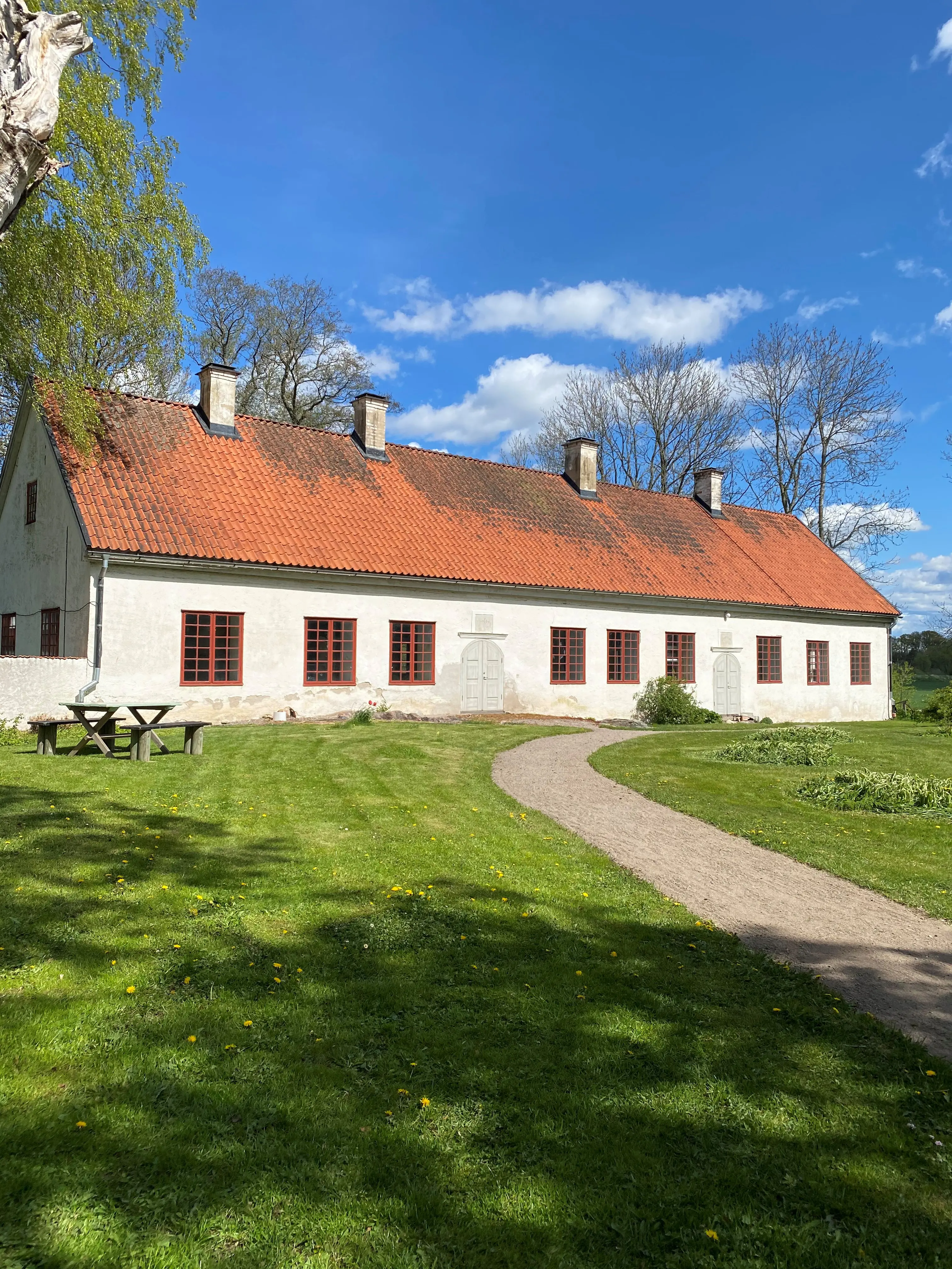 Lilla Stenhuset på Tuna kungsgård. En vitputsad byggnad med brant tegeltak, stora, spröjsade fönster och två rundade portar. Vår och skir grönska.