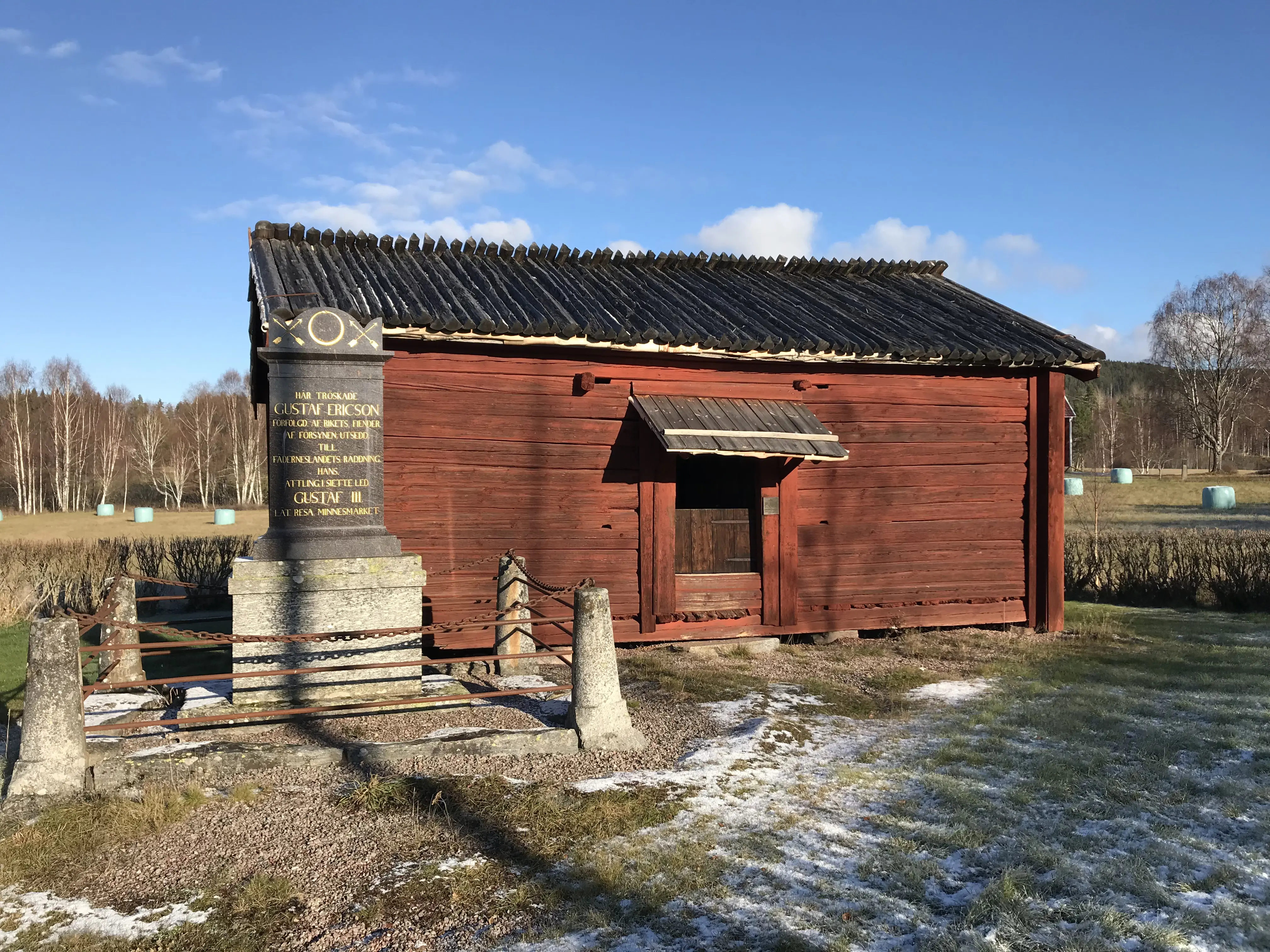 Isala kungslada. En liten, rödmålad byggnad i liggande timmer. Framför ladan står ett pampigt stenmonument över Gustav Vasa. Tidig vår.