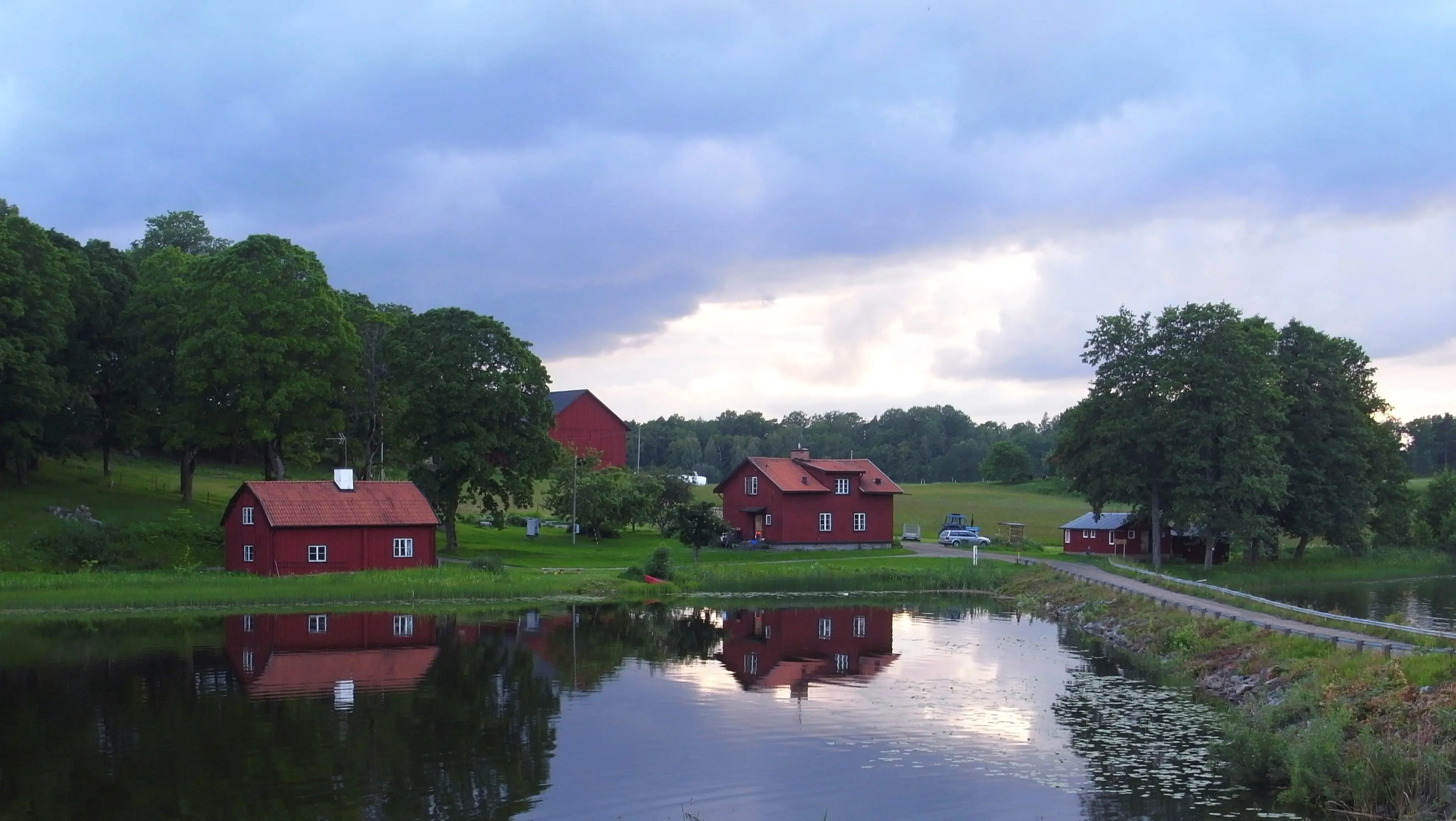 Ådö kaptensboställe sett över vattnet. Ett par röda byggnader speglas i sjön Båven. Skymning, sommar och grönska.