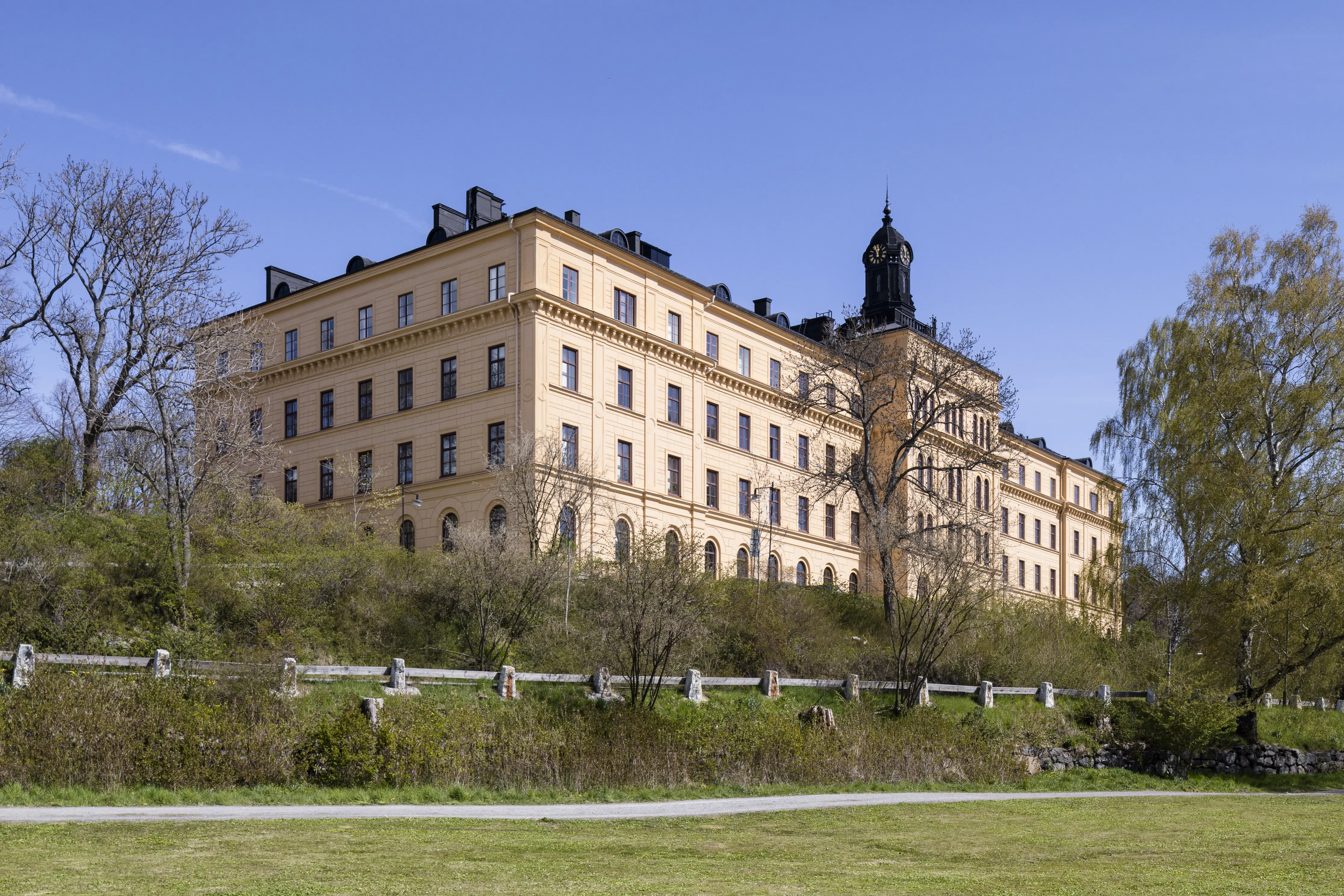 Manillaskolans huvudbyggnad sedd nedifrån strandpromenaden på Djurgården. En massiv byggnad i gul puts med lanternin på taket.